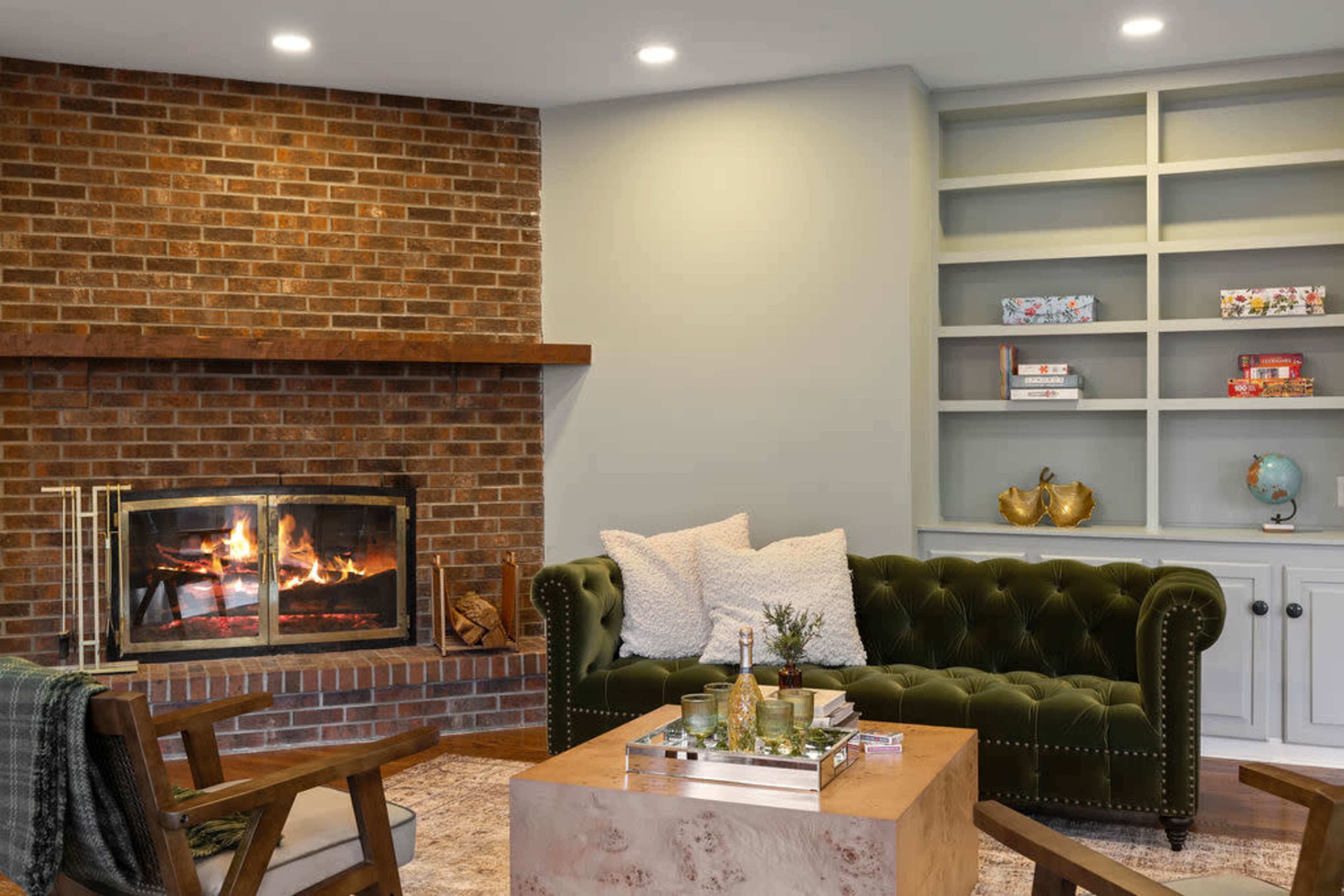 A cozy living room features a brick fireplace, a green velvet sofa with decorative pillows, a wooden coffee table, and shelves with various decorative items.