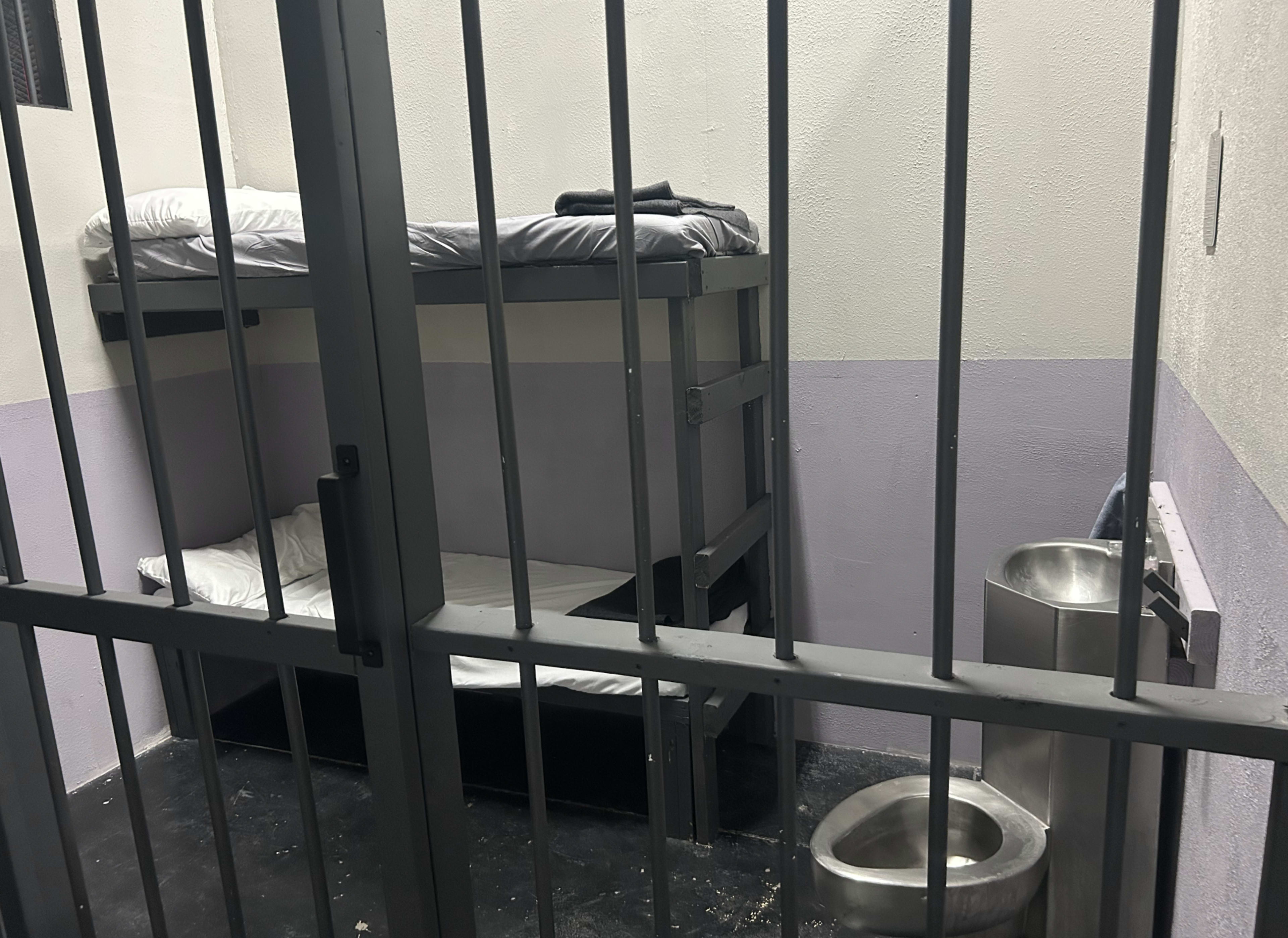 The image shows a small jail cell with two metal bunk beds and a toilet adjacent to the bars.