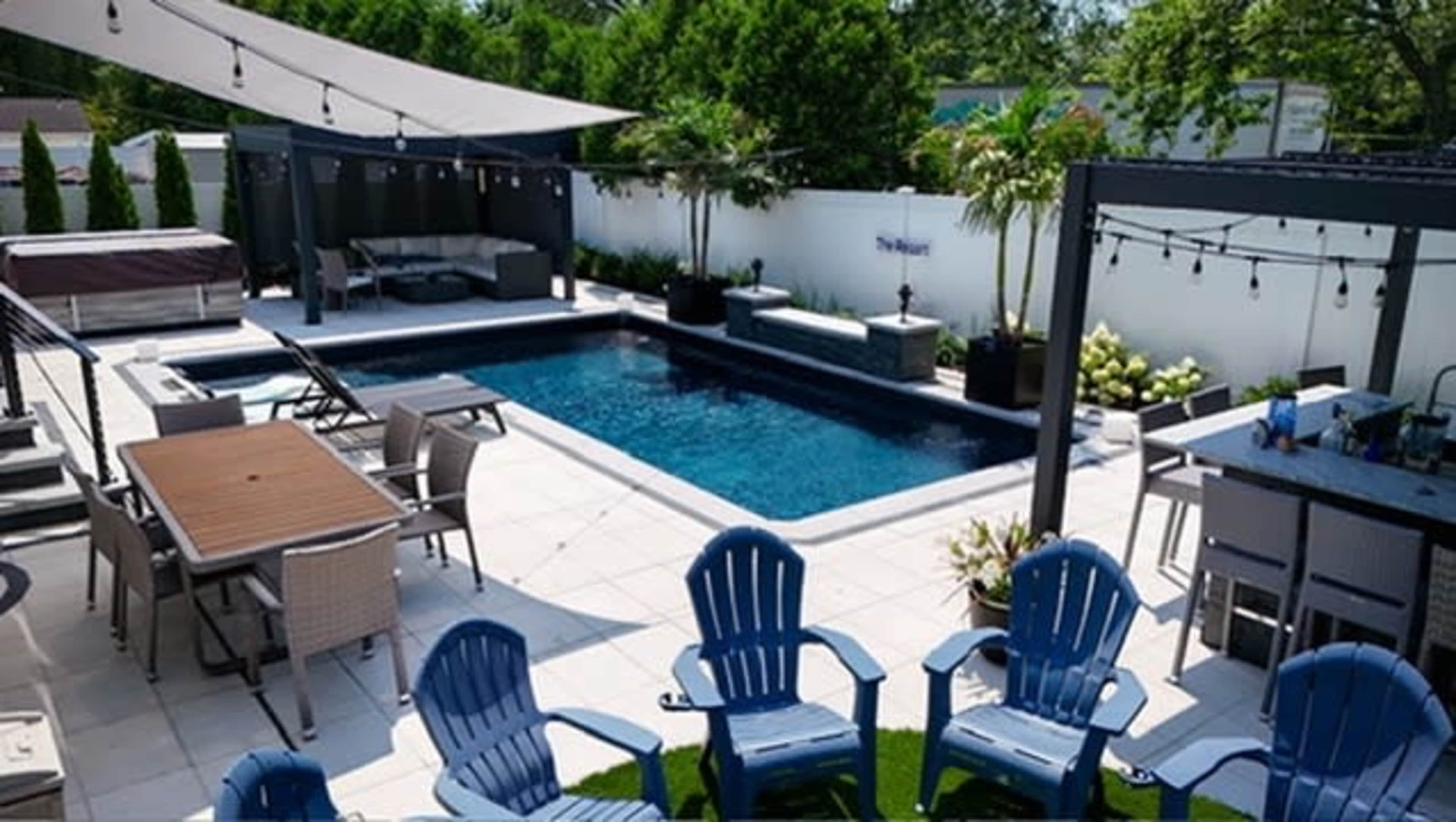 The image shows a landscaped backyard with a swimming pool, lounge chairs, outdoor seating, and a bar area under shaded structures.