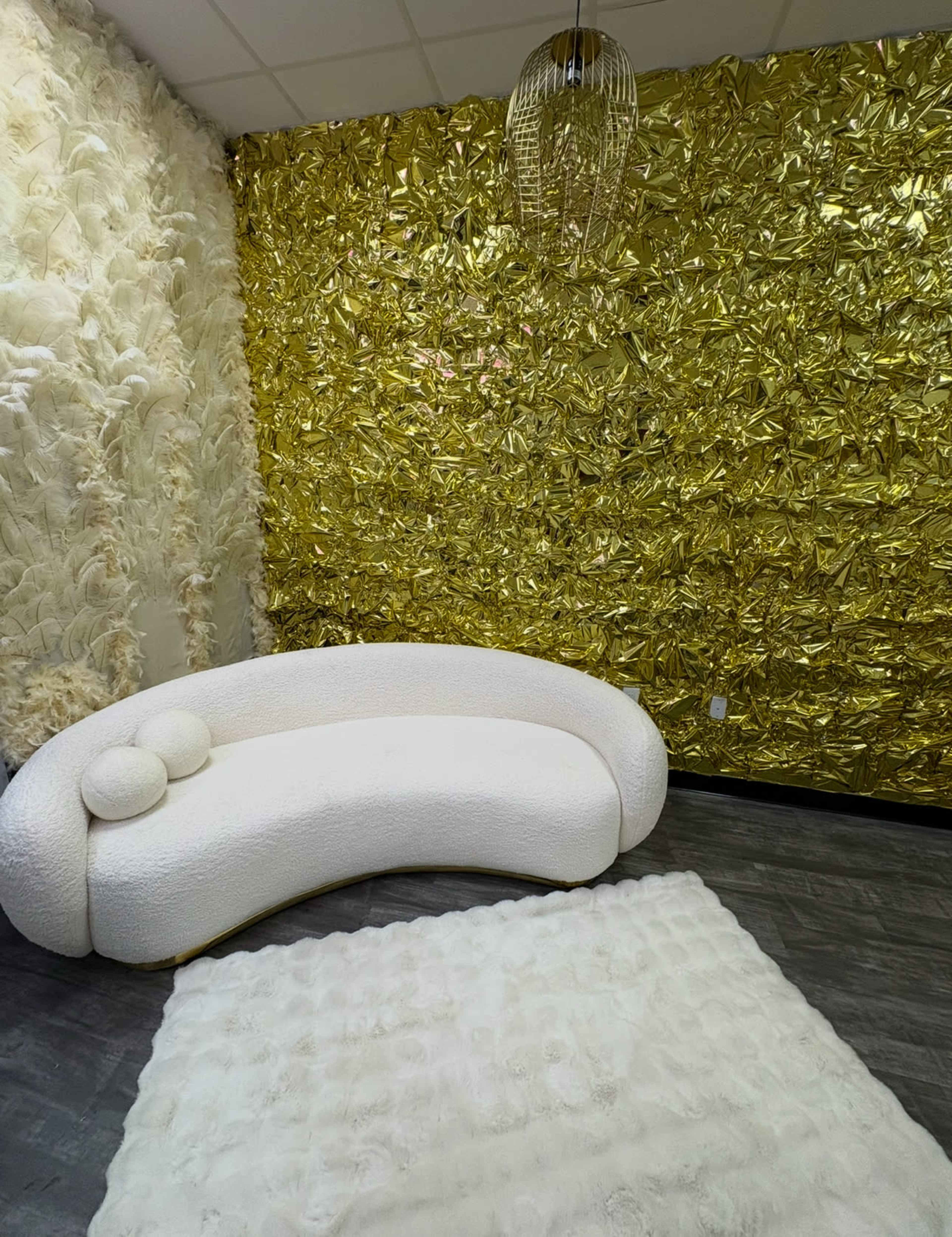 The image shows a small room featuring a curved white sofa with two round cushions, a textured white rug on the floor, and one wall covered in shiny gold foil.