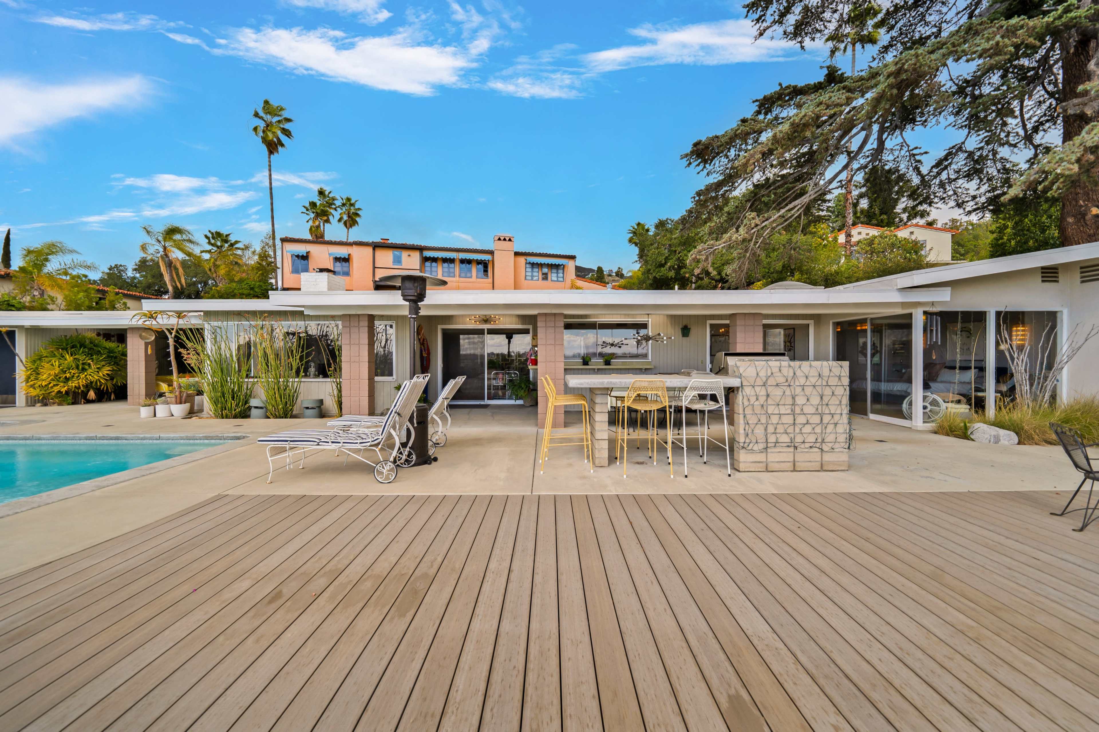 The image shows a modern outdoor patio area with a swimming pool, surrounded by lounge chairs, a bar table, and a backdrop of a residential building and palm trees.