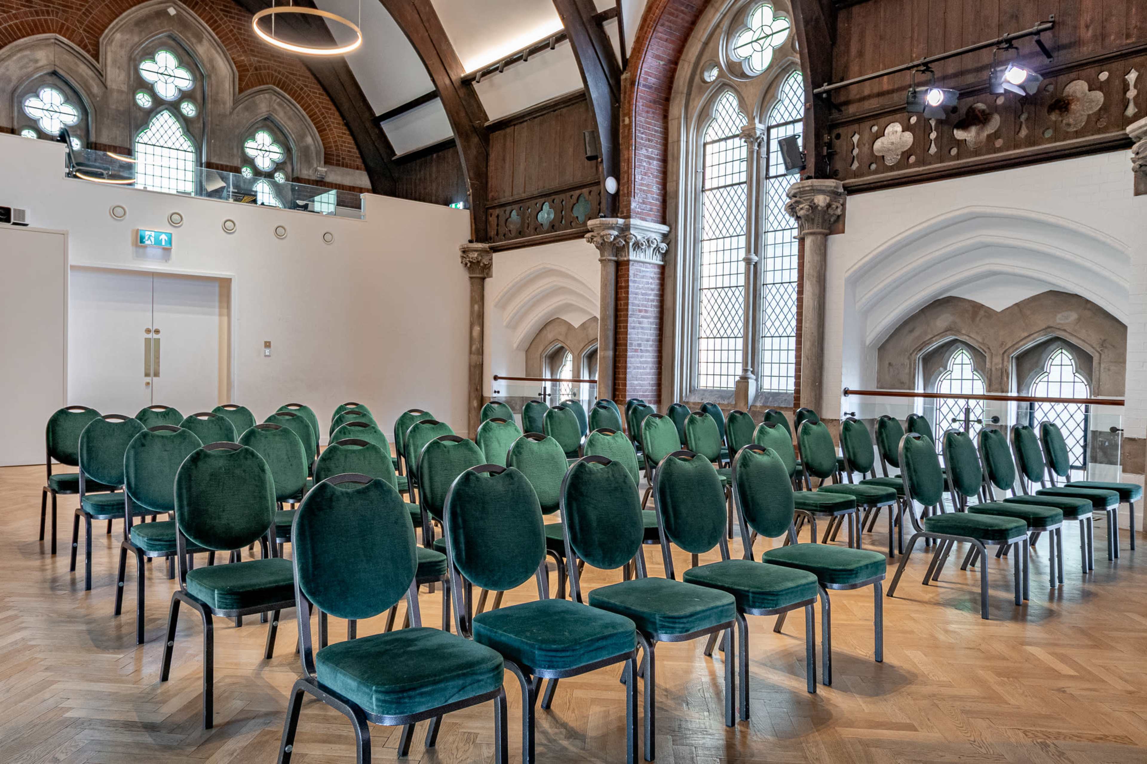 A row of green velvet chairs is arranged in a formal seating layout inside a large, well-lit hall with arched windows and wooden beams.