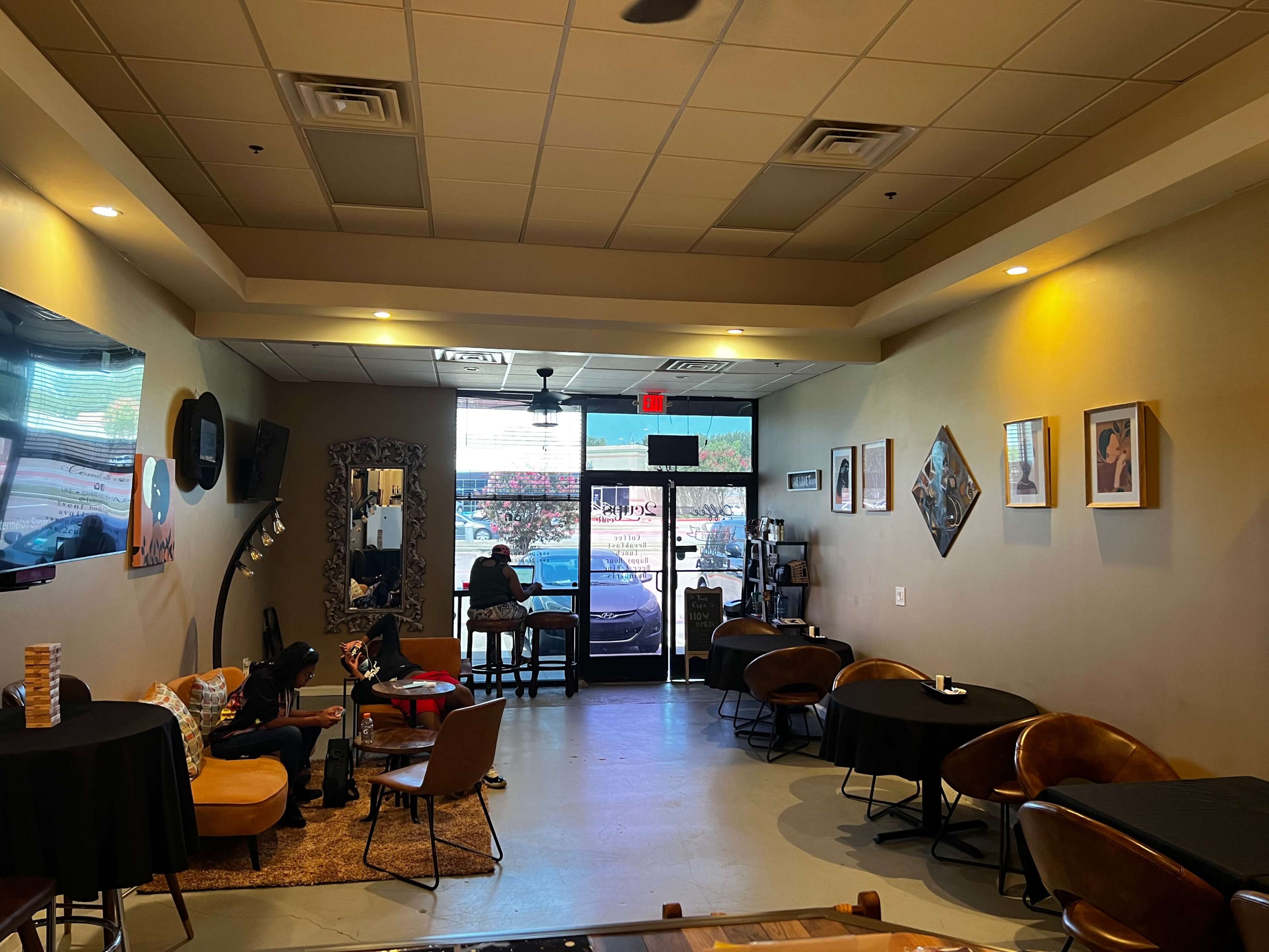 A cozy café interior with several tables and chairs, featuring a customer seated near the entrance and natural light streaming through the front window.