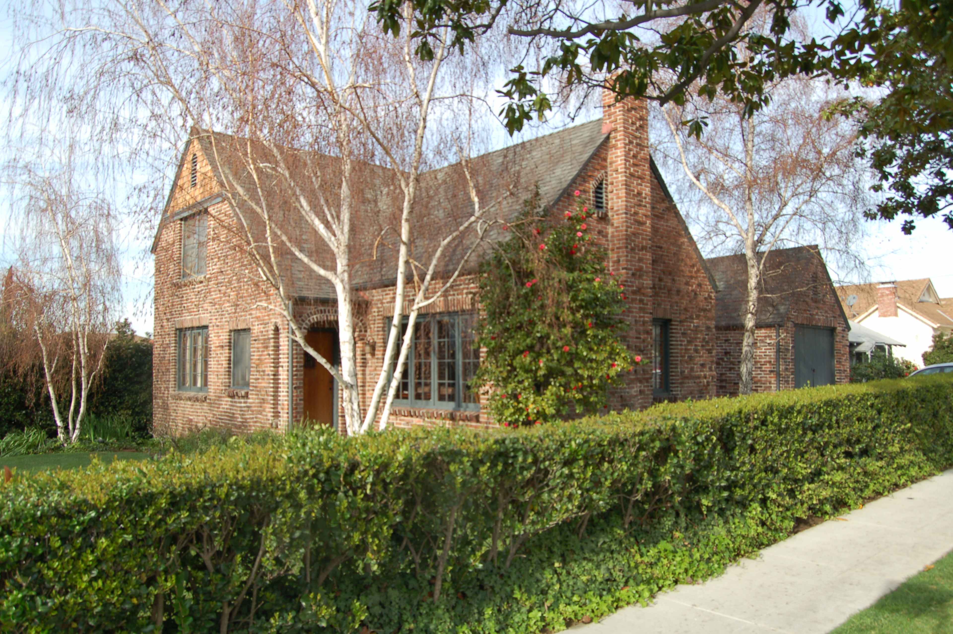 A brick house with a steep roof is surrounded by a neatly trimmed hedge and several birch trees.