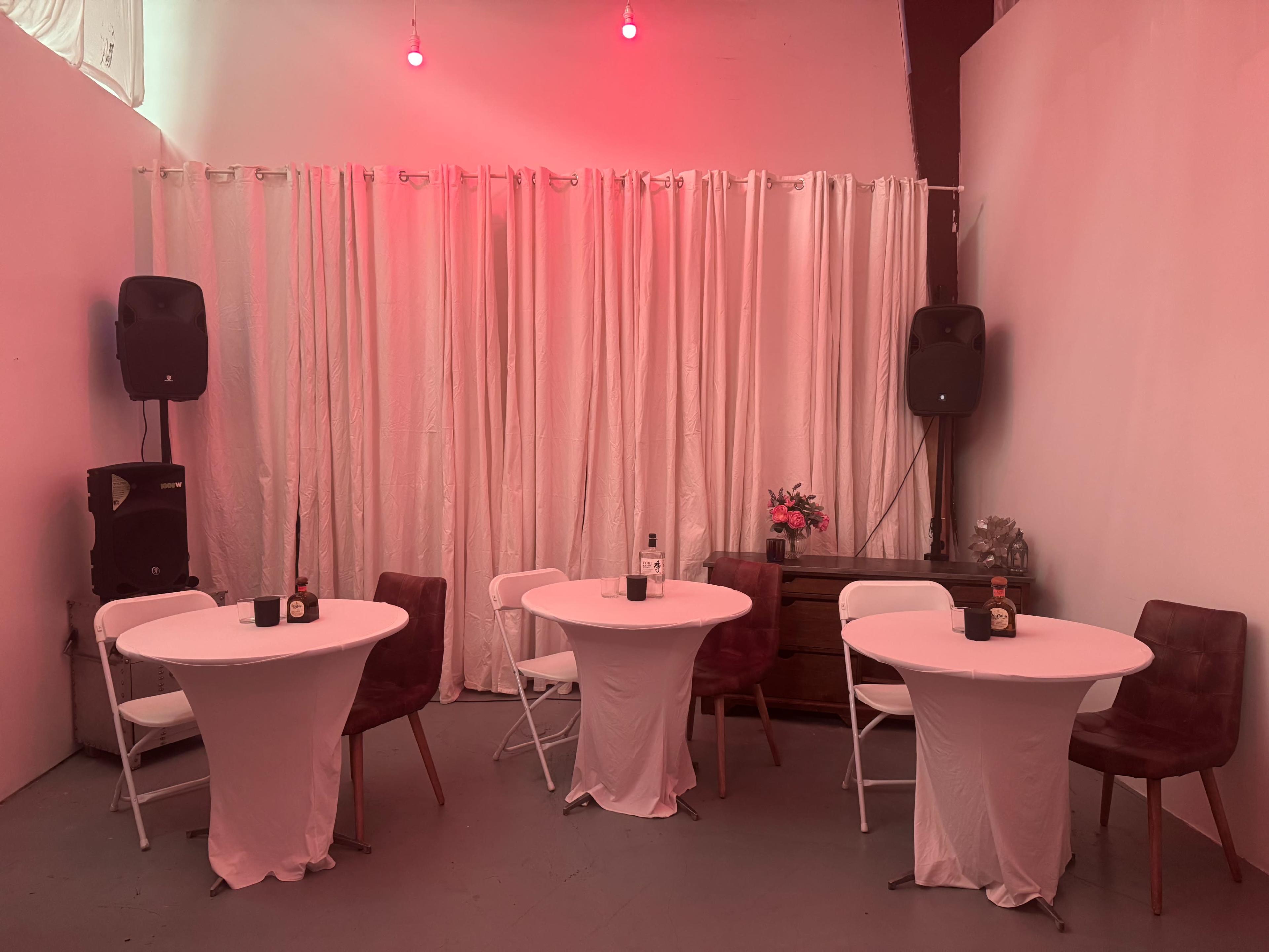 The image shows a small event space with two round tables covered in white cloth, surrounded by four chairs, a wooden dresser, and pink-lit curtains in the background.