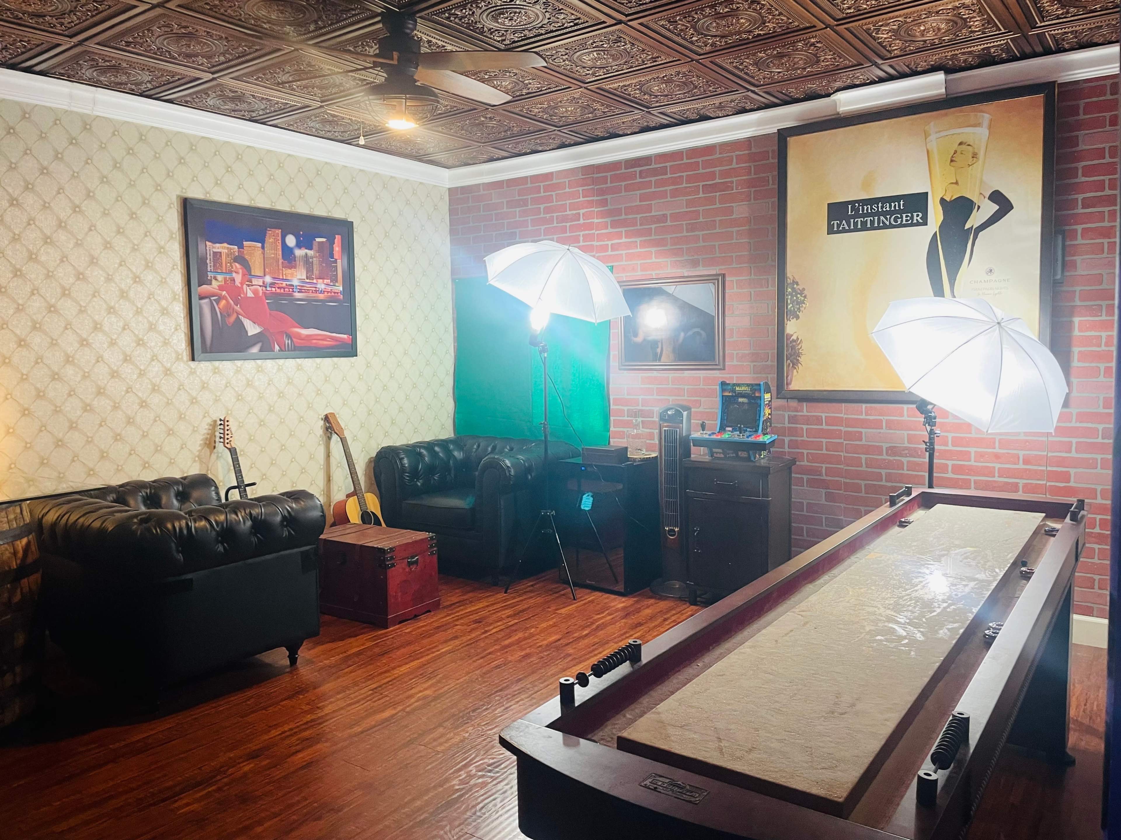 The room features a shuffleboard table, two couches, a vintage-style poster, and studio lighting equipment.