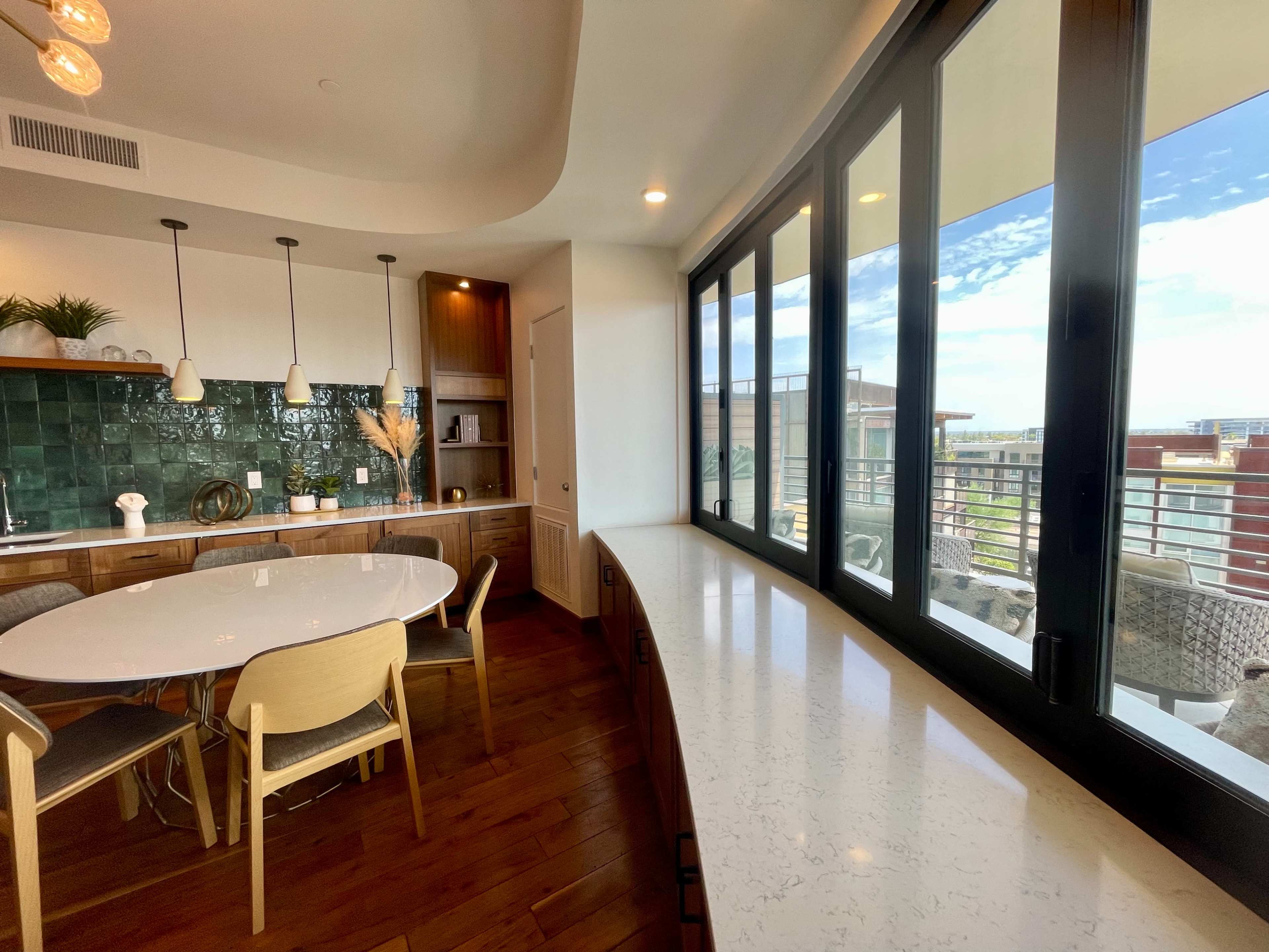 The image shows a modern dining area with a circular table surrounded by chairs and large windows offering a view of the outdoors.