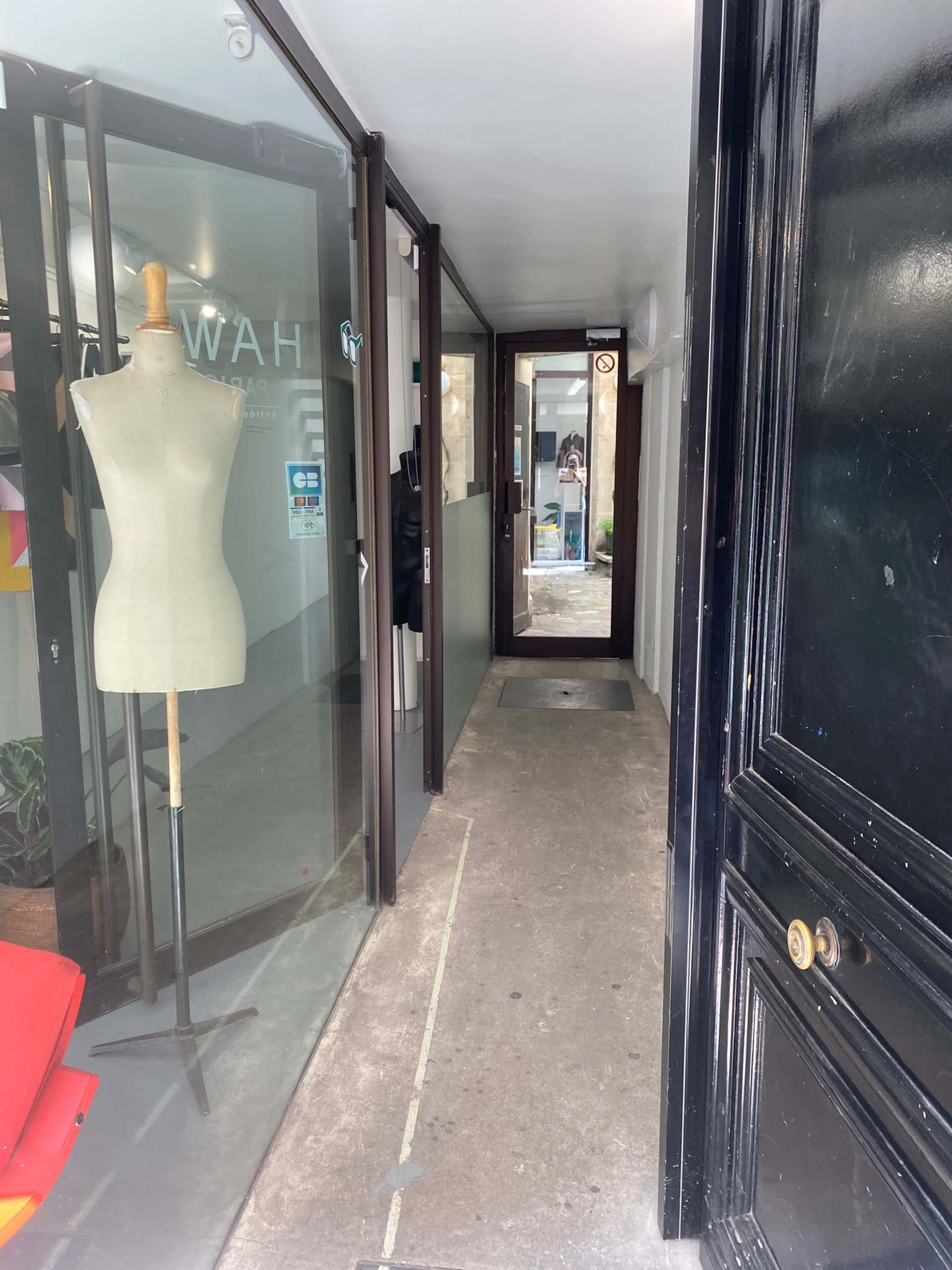 The image shows a narrow hallway with glass walls, a dress form on the left, and an entrance at the far end.