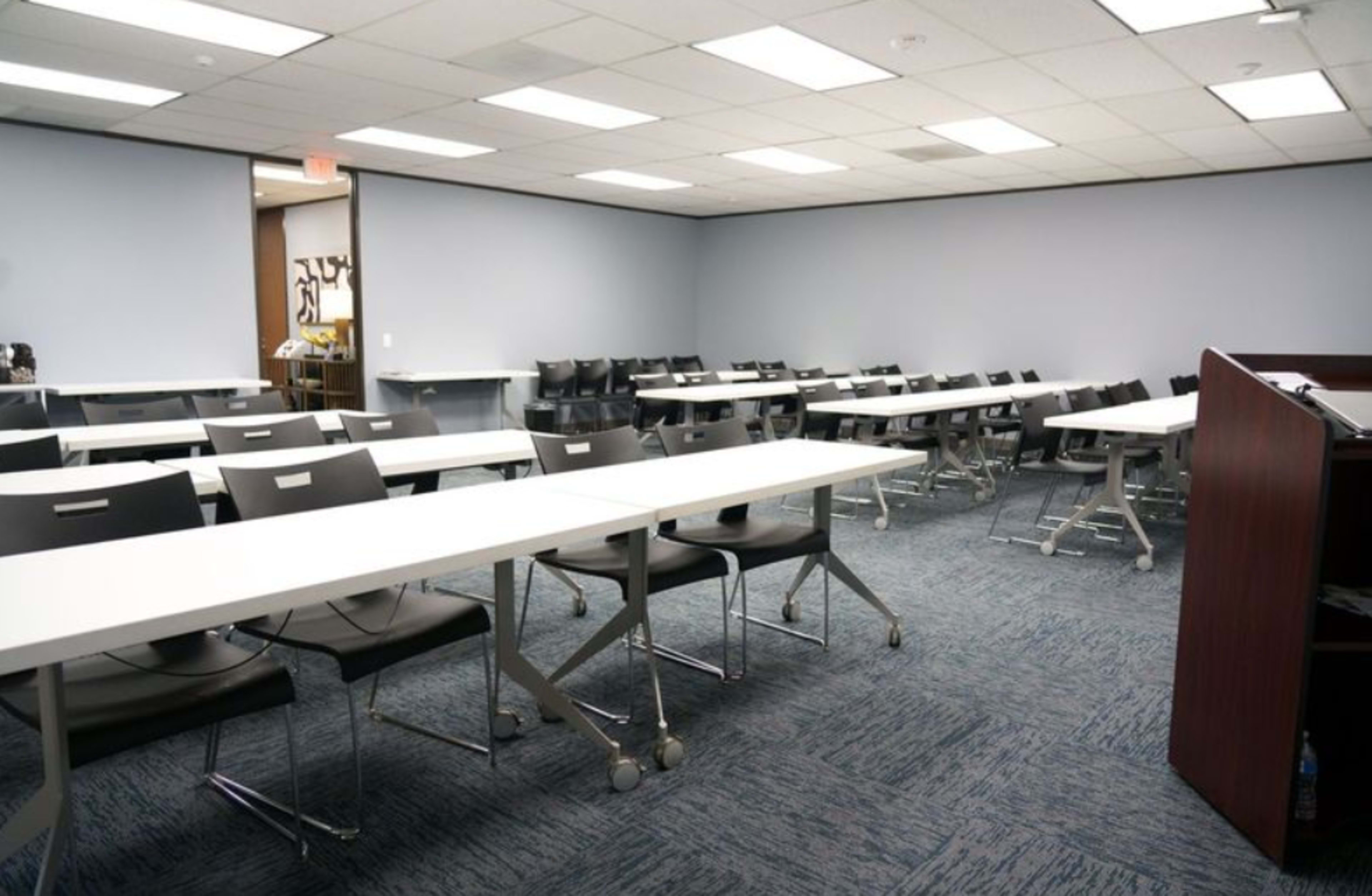The image shows a modern classroom with several long tables arranged in rows and black chairs positioned around them.