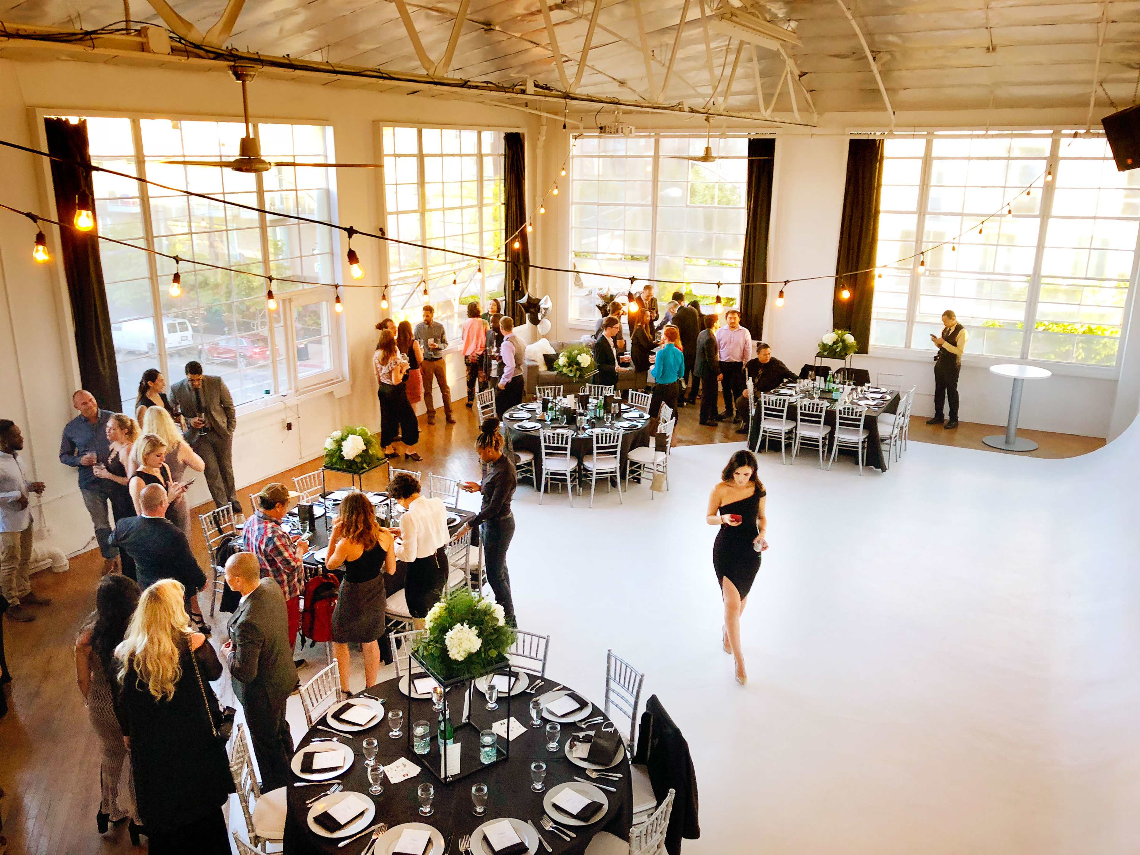 A spacious event venue is filled with people socializing at elegantly set tables, while a woman in a black dress walks across the floor.