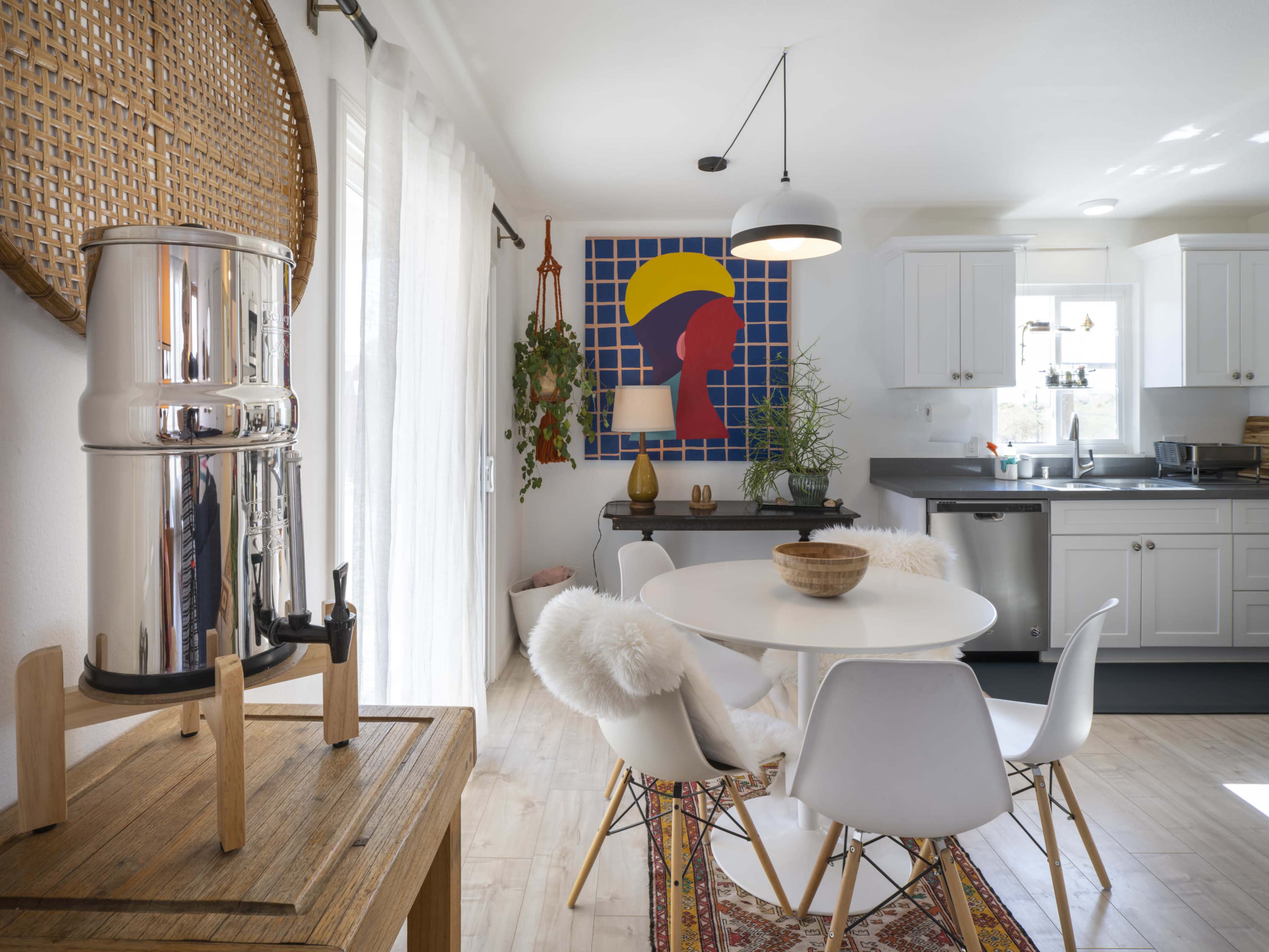 A modern kitchen and dining area features a round table with white chairs, a stainless steel water filter, and a colorful wall art piece.