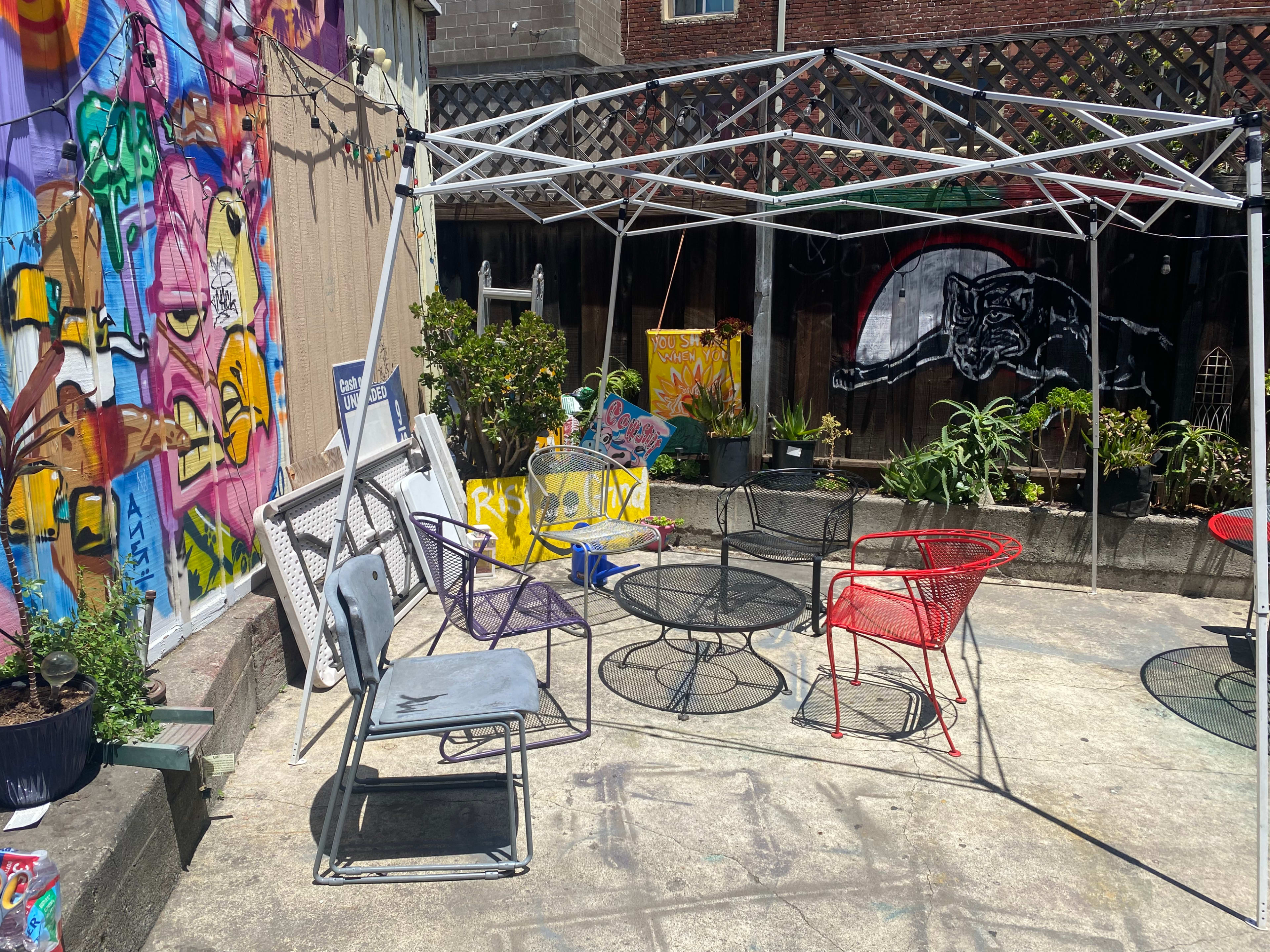 A shaded seating area with various chairs and tables is set up beside a colorful mural and potted plants.