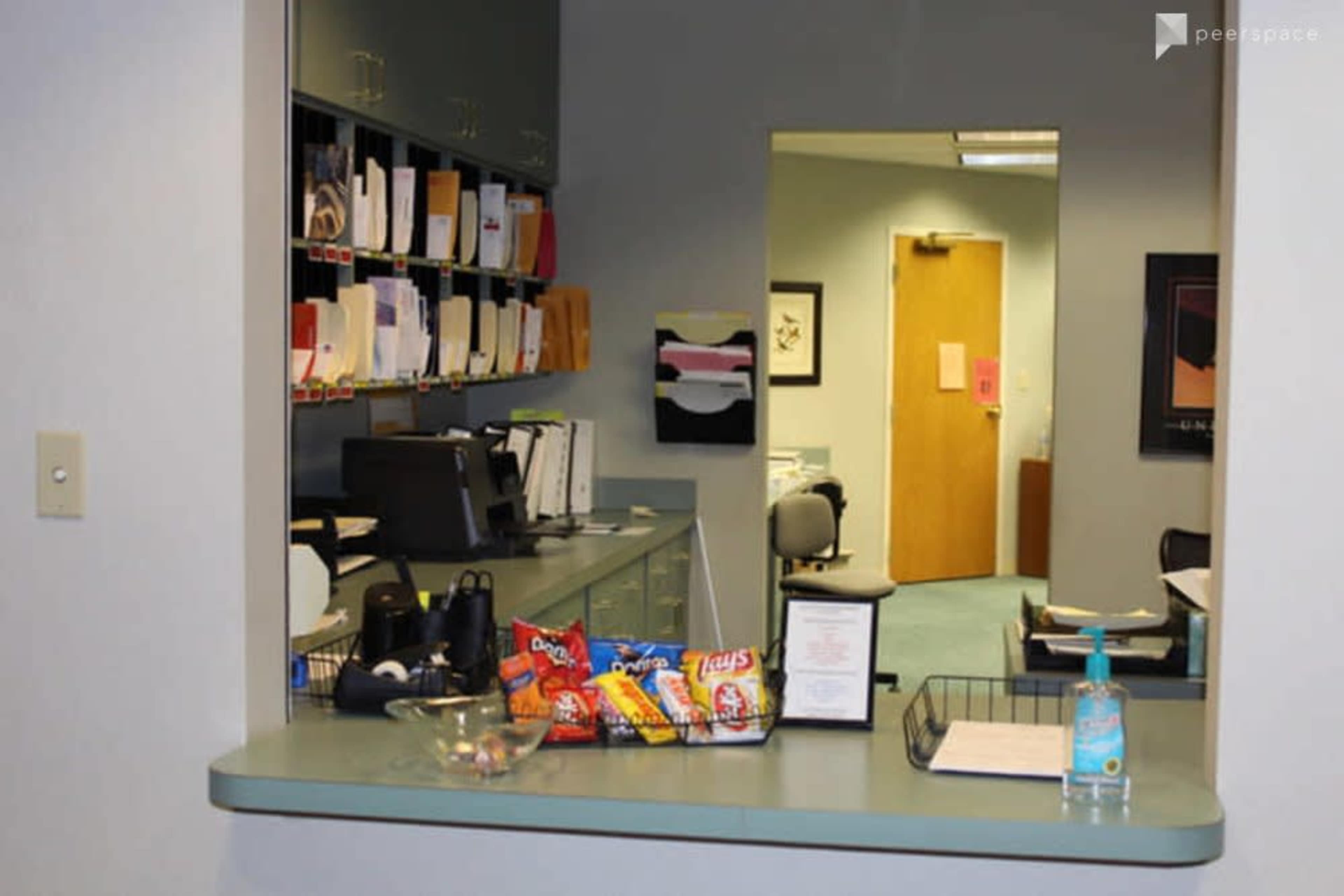 The image shows an office space with a reception area featuring a desk, files on the wall, and a snack display with various chips.