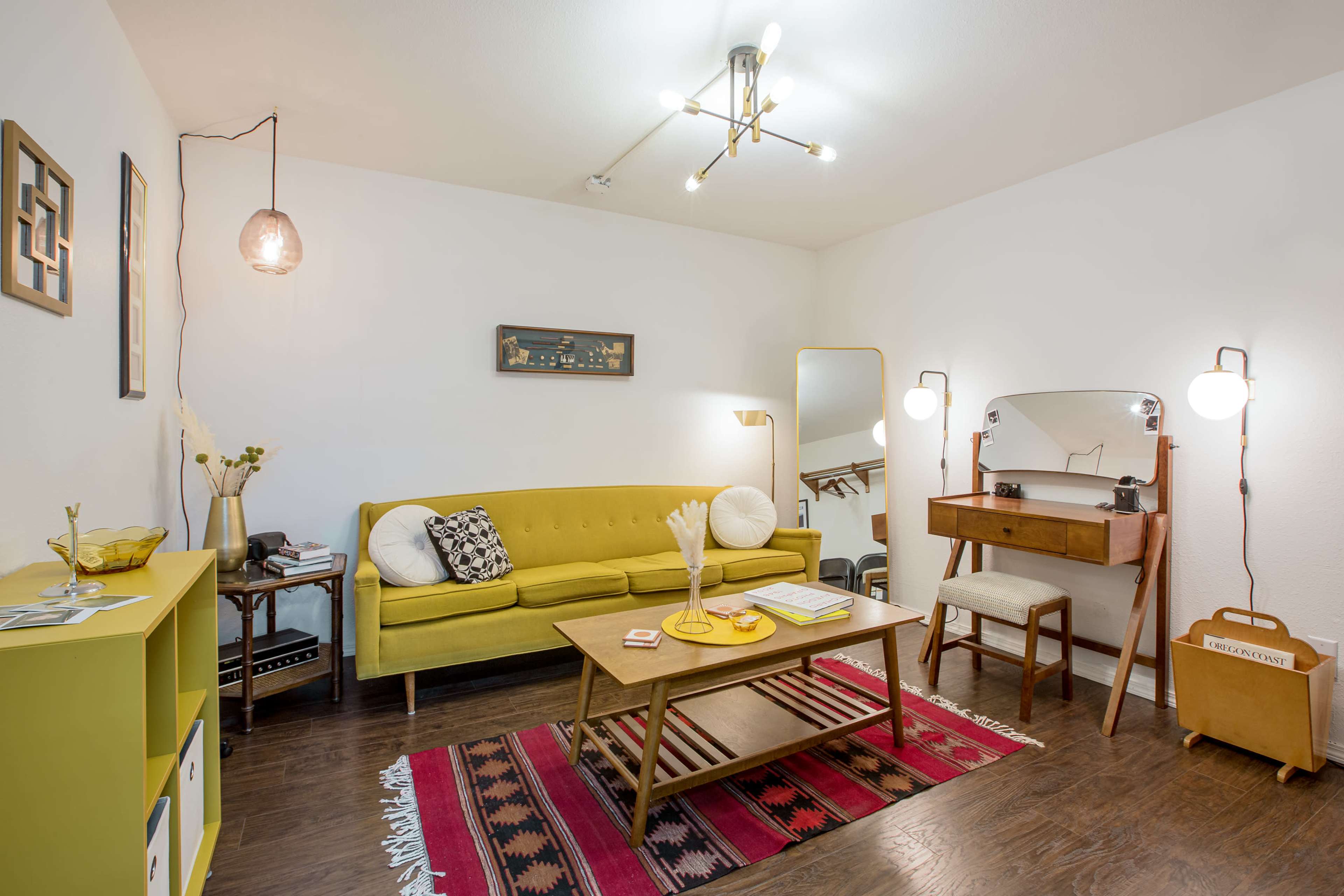 A cozy living room featuring a yellow sofa, a wooden coffee table on a patterned rug, and a dressing table against a white wall.