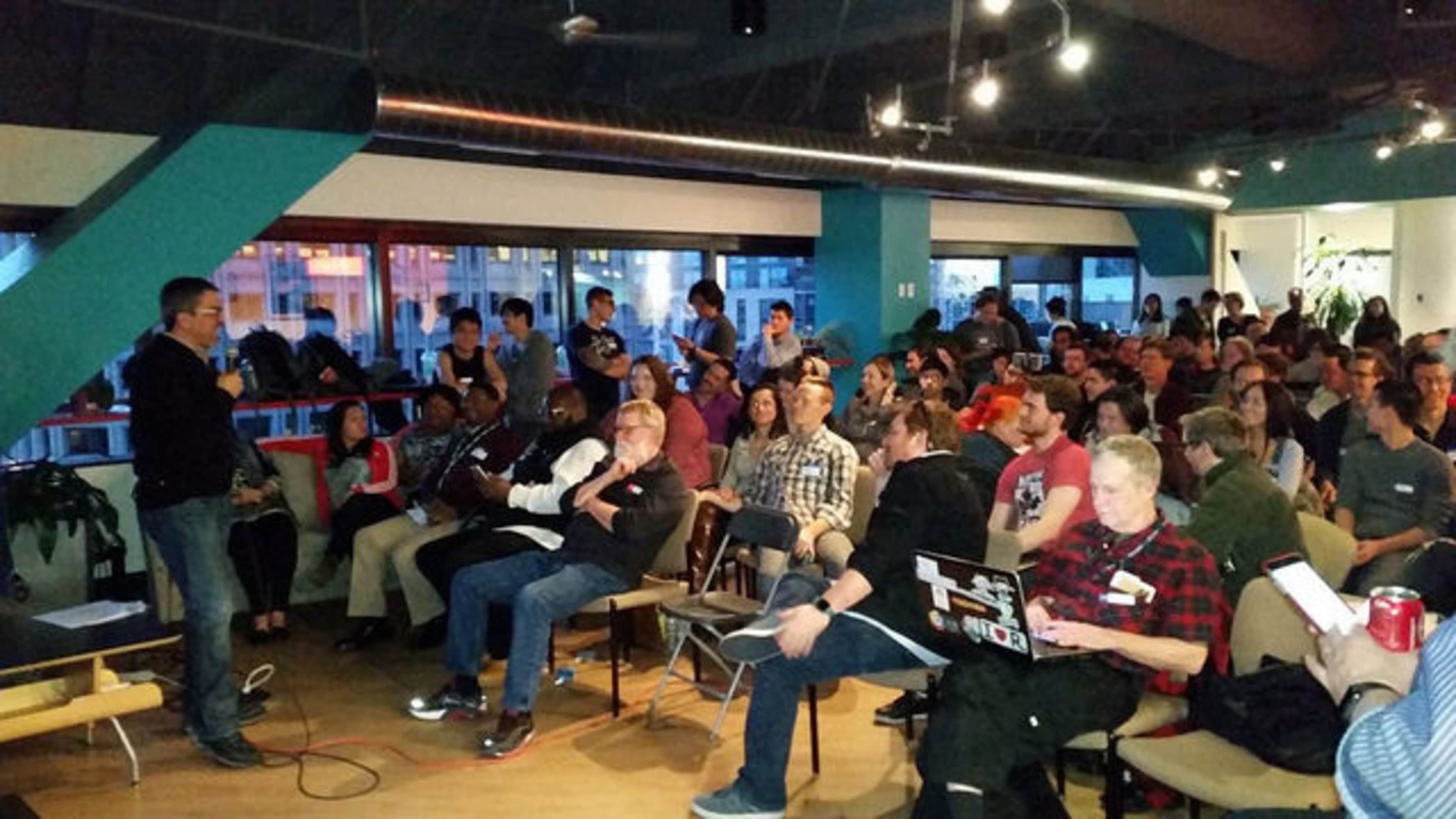 A large group of people is seated in a modern room, attentively watching a speaker at the front of the space.