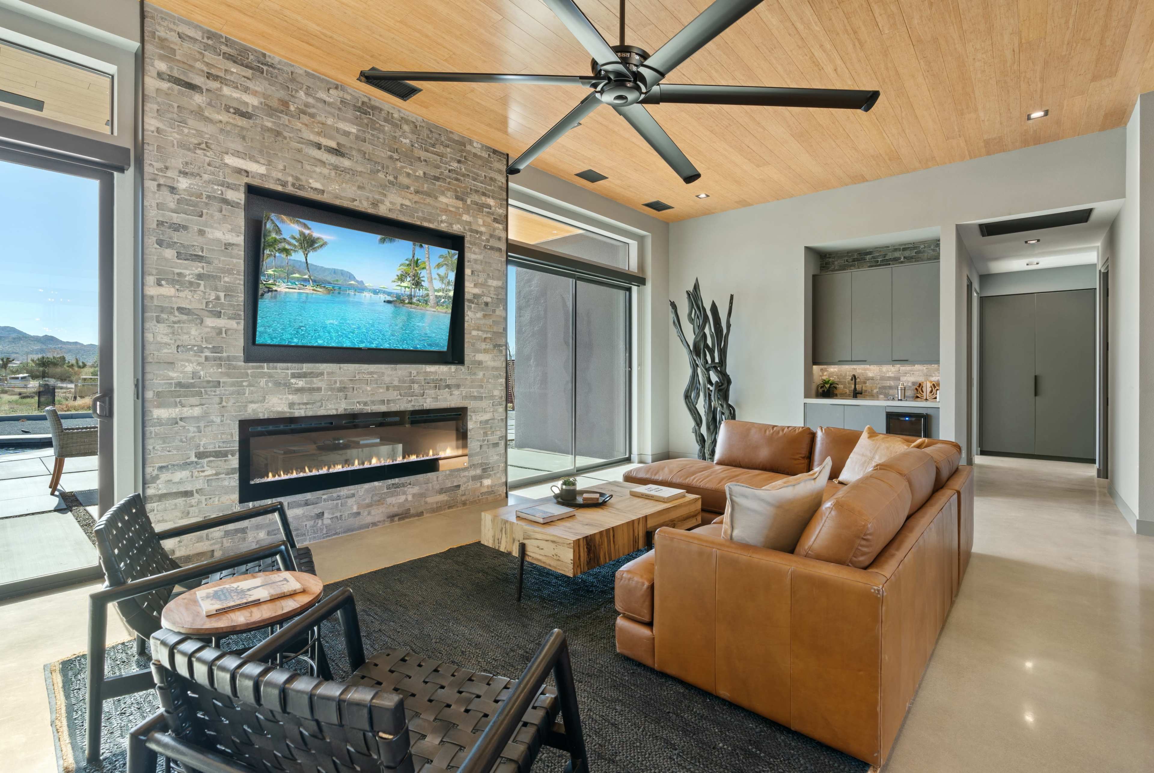 The living room features a leather sofa, modern art, a stone wall with a television, and a fireplace, all illuminated by large windows.