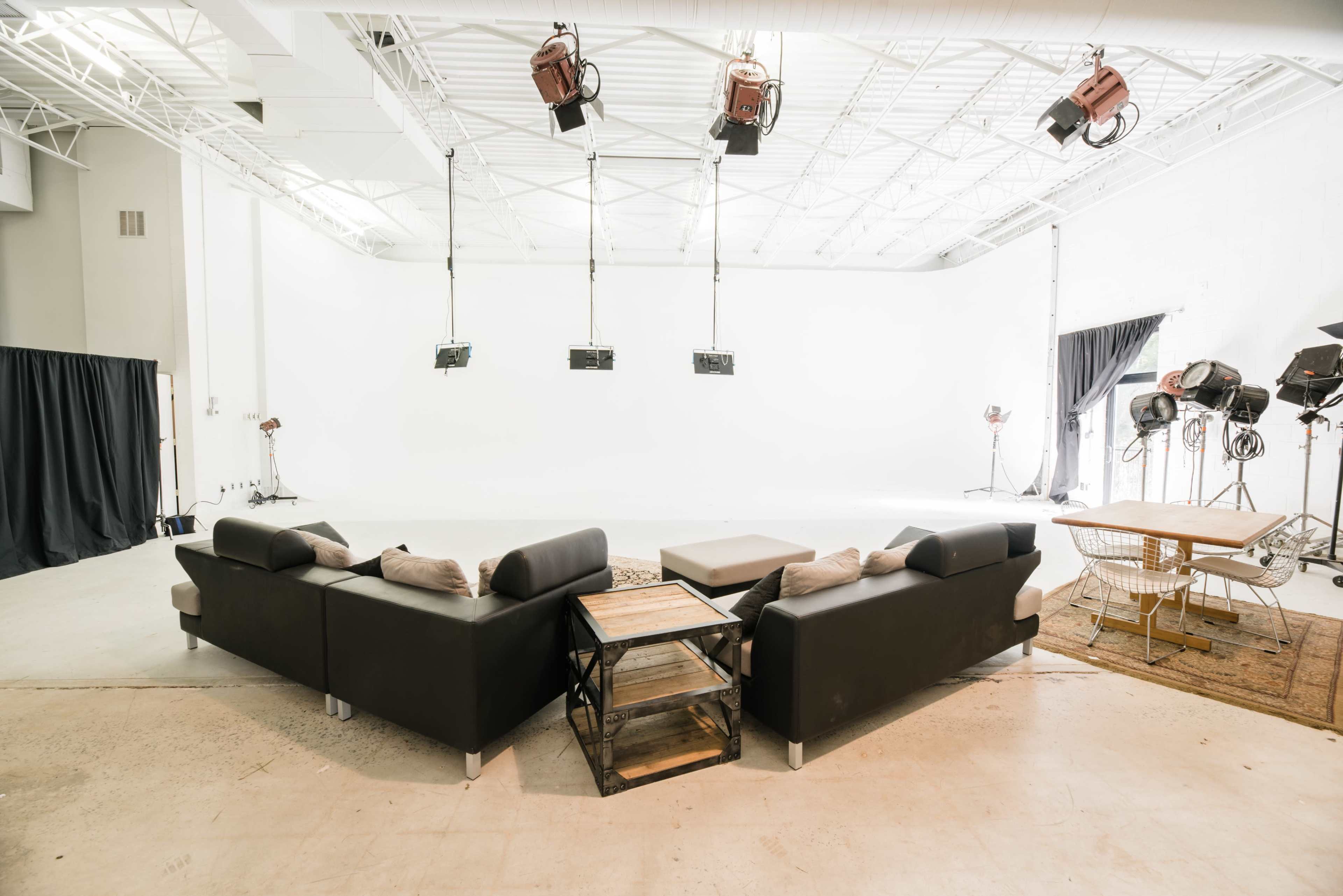 A spacious indoor studio with a black sectional sofa, a wooden coffee table, and several studio lights overhead.
