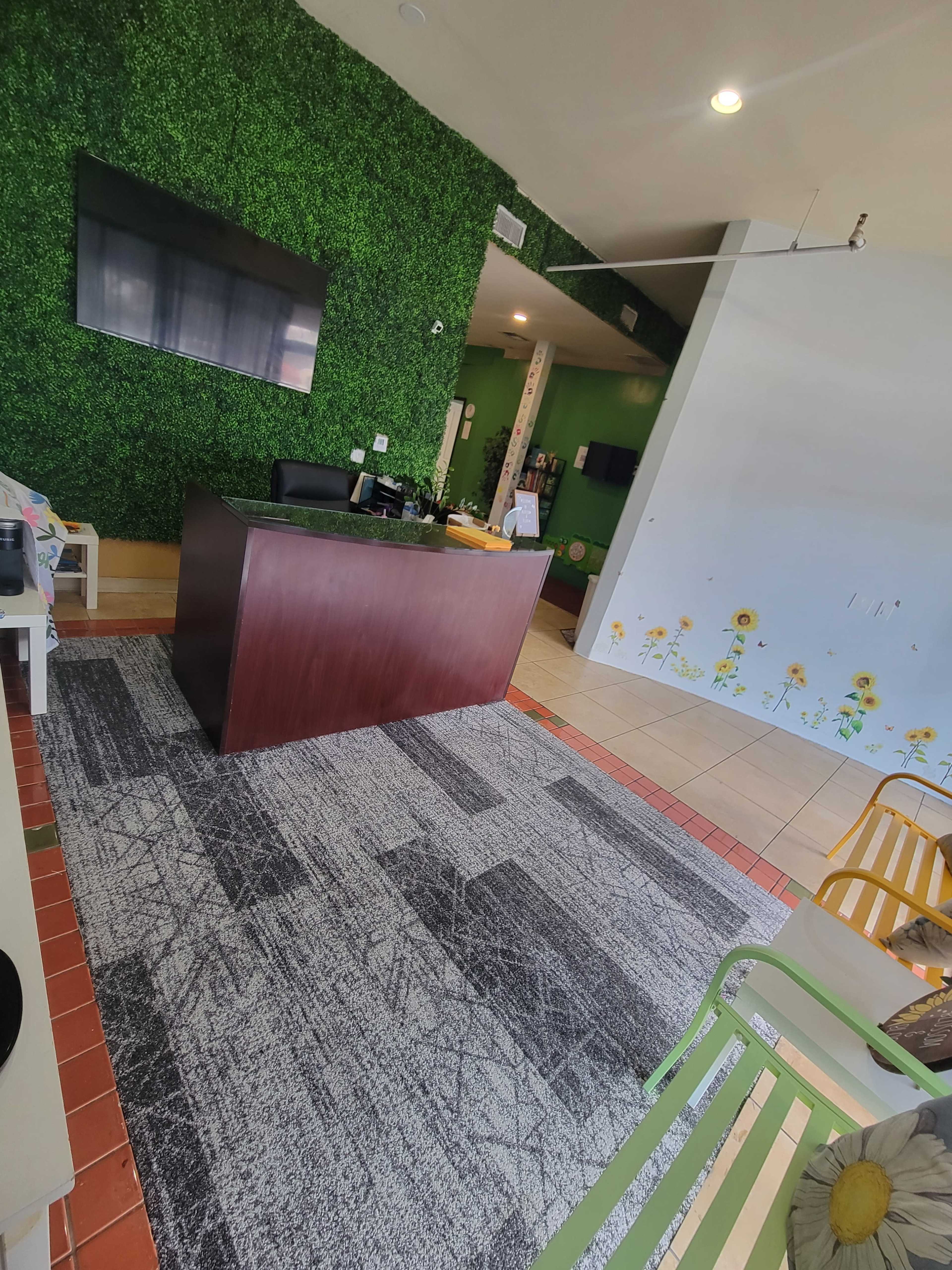 The image shows a reception area with a wooden desk, green wall decor featuring artificial plants, and a large television mounted on the wall.