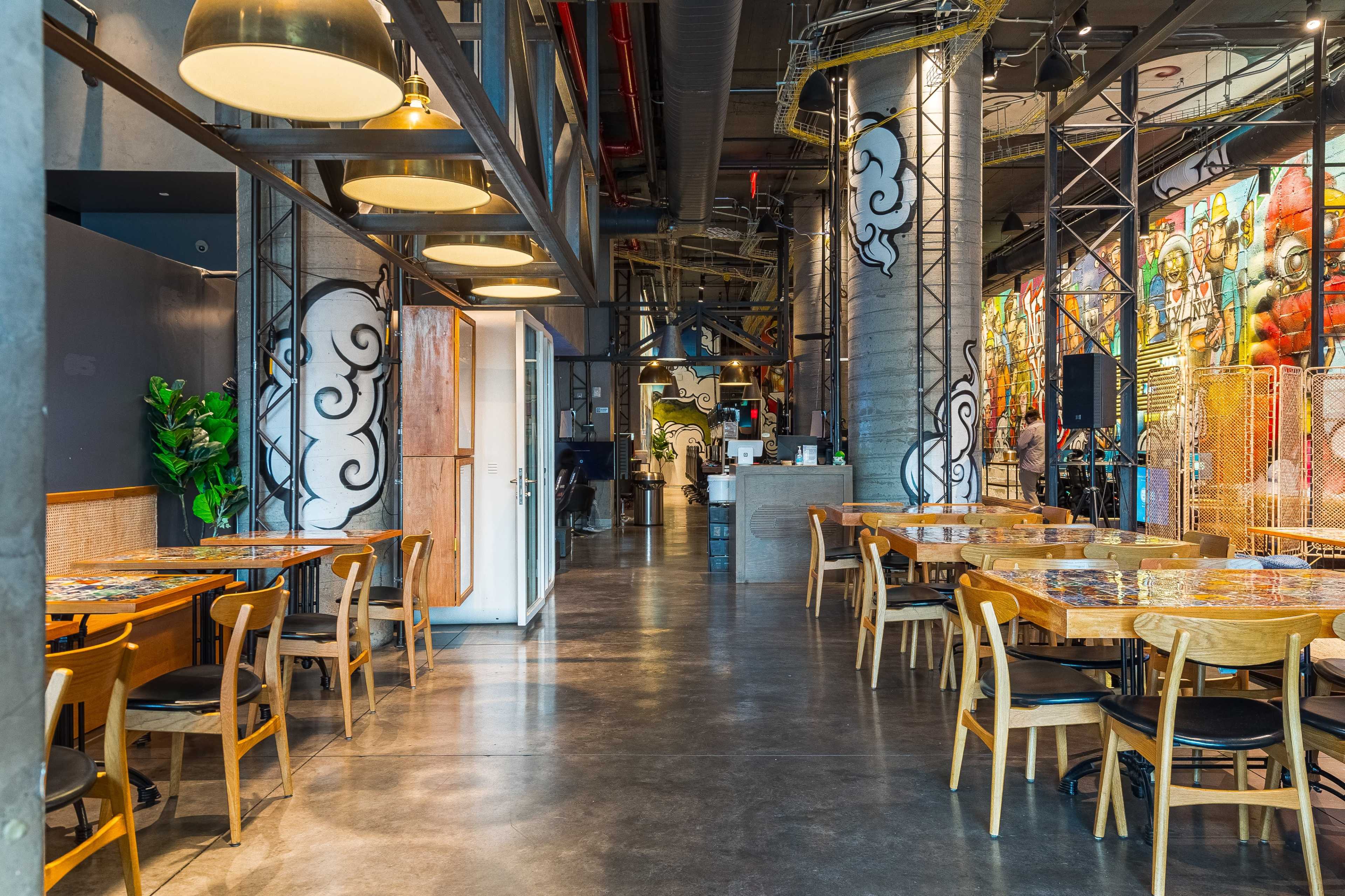 A modern dining area with wooden tables and chairs, industrial-style decor, and vibrant murals on the walls.