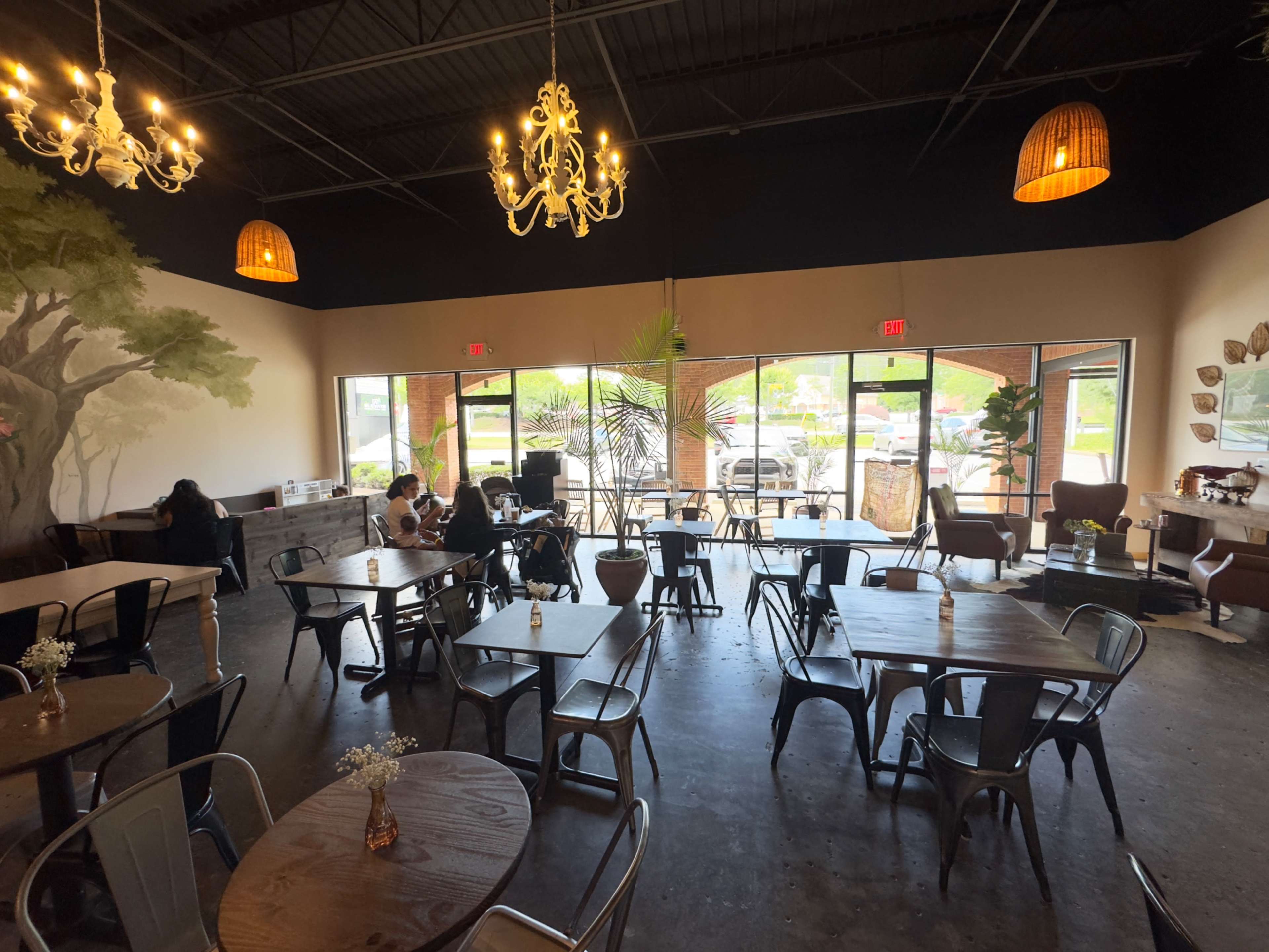 The image shows a spacious café with wooden tables, metal chairs, and large windows that provide natural light, featuring a mural on one wall and patrons seated at tables.