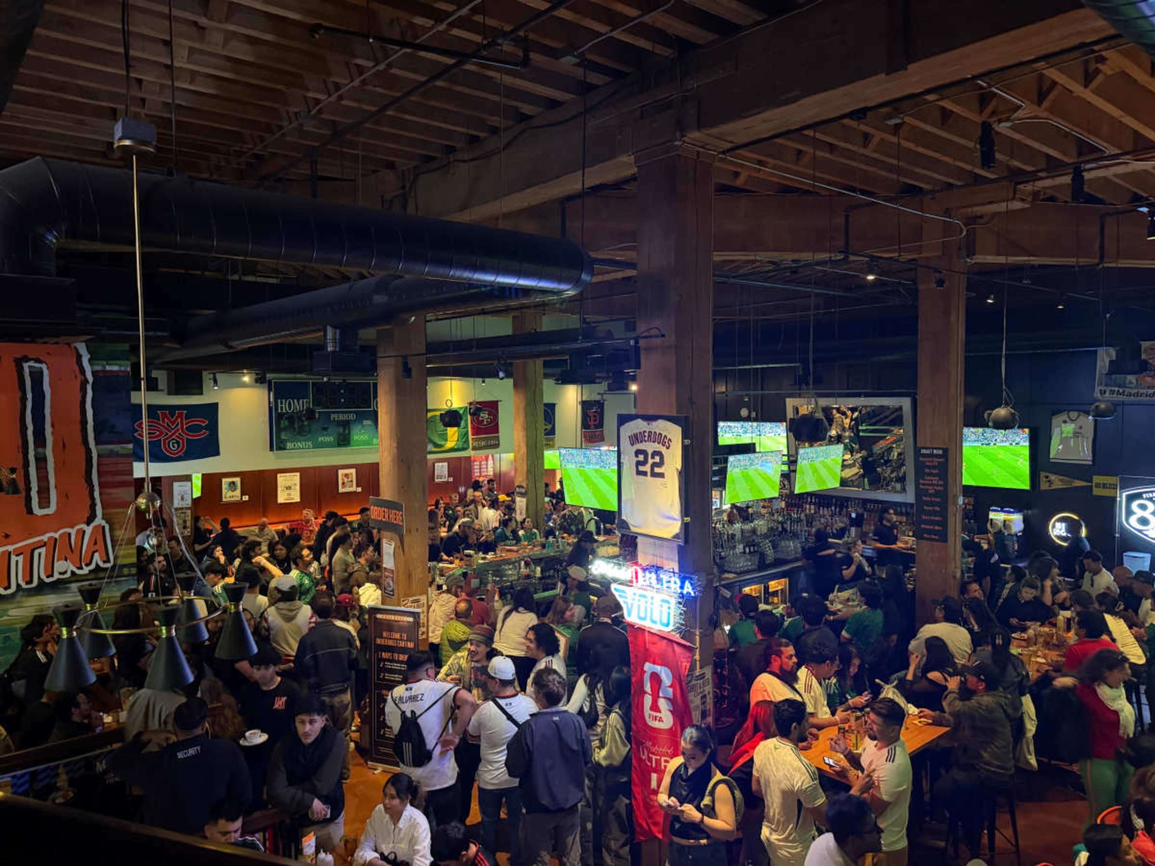 A crowded sports bar features multiple large screens displaying a soccer match while patrons eat and socialize at tables.