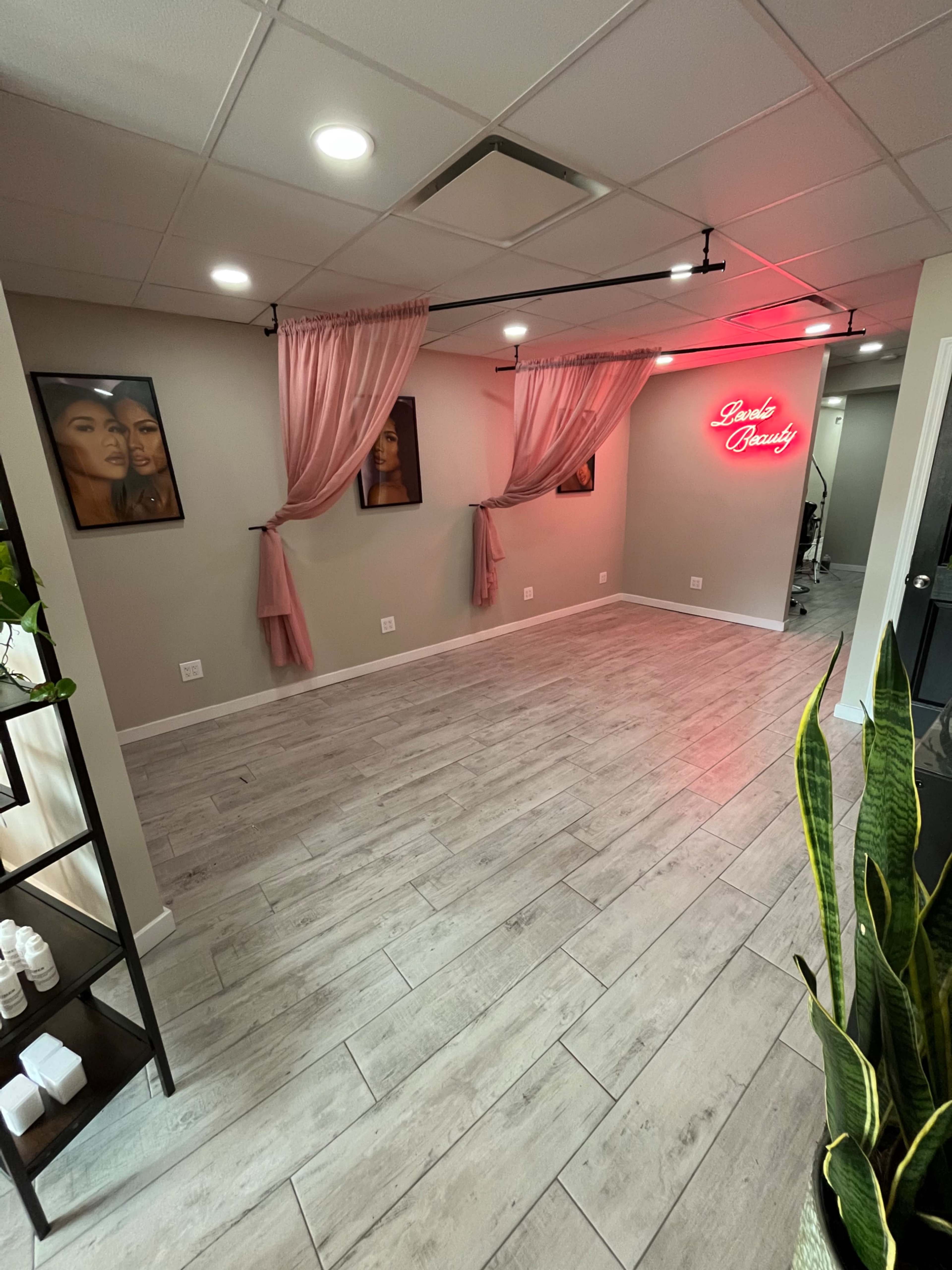 A beauty salon interior with light-colored wooden flooring, pink curtains, and a neon sign reading "Dani's Beauty" on the wall, alongside framed portraits of two women.