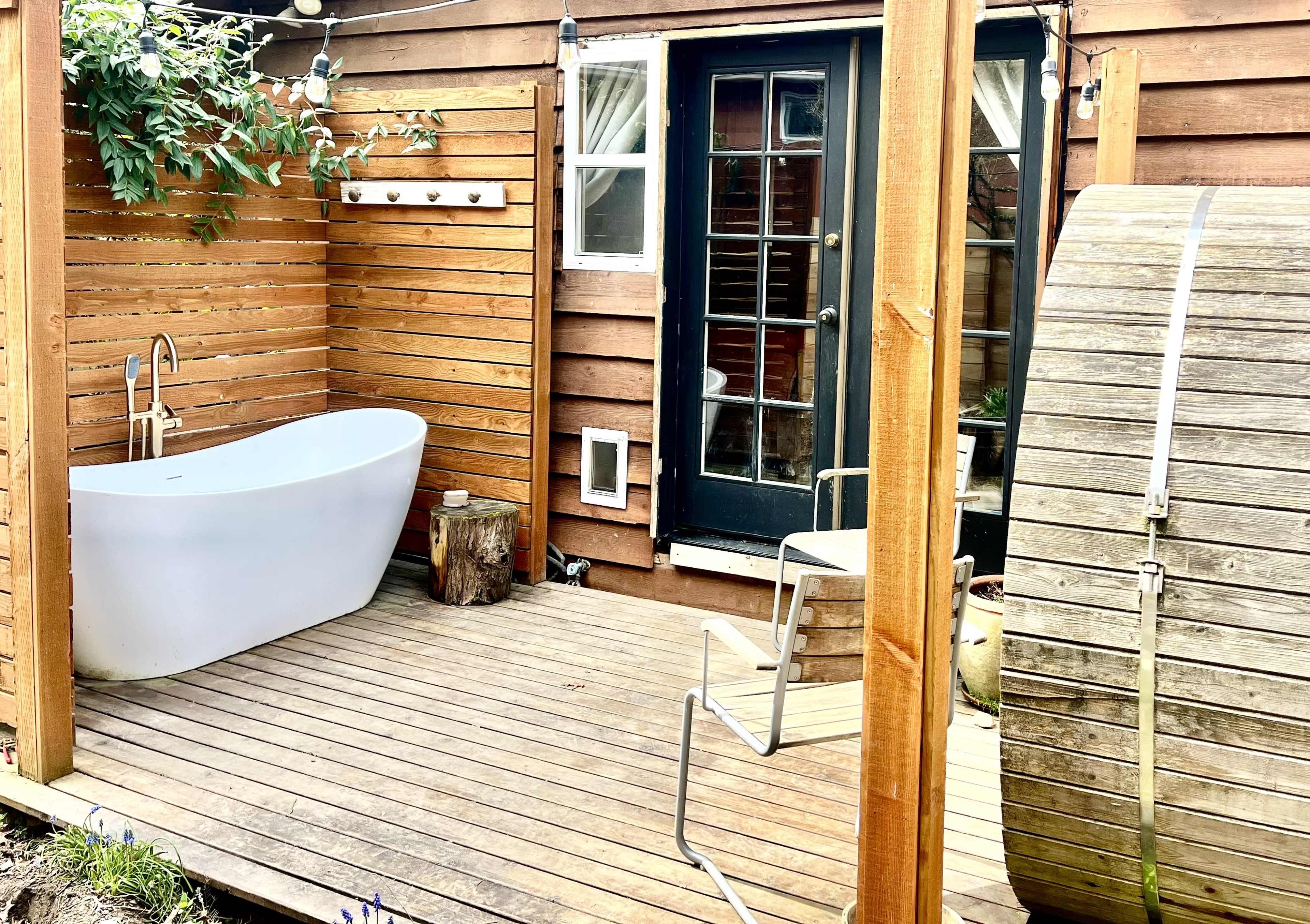 The image shows a wooden deck area featuring a freestanding white bathtub, a wooden barrel, and a door leading to an interior space.