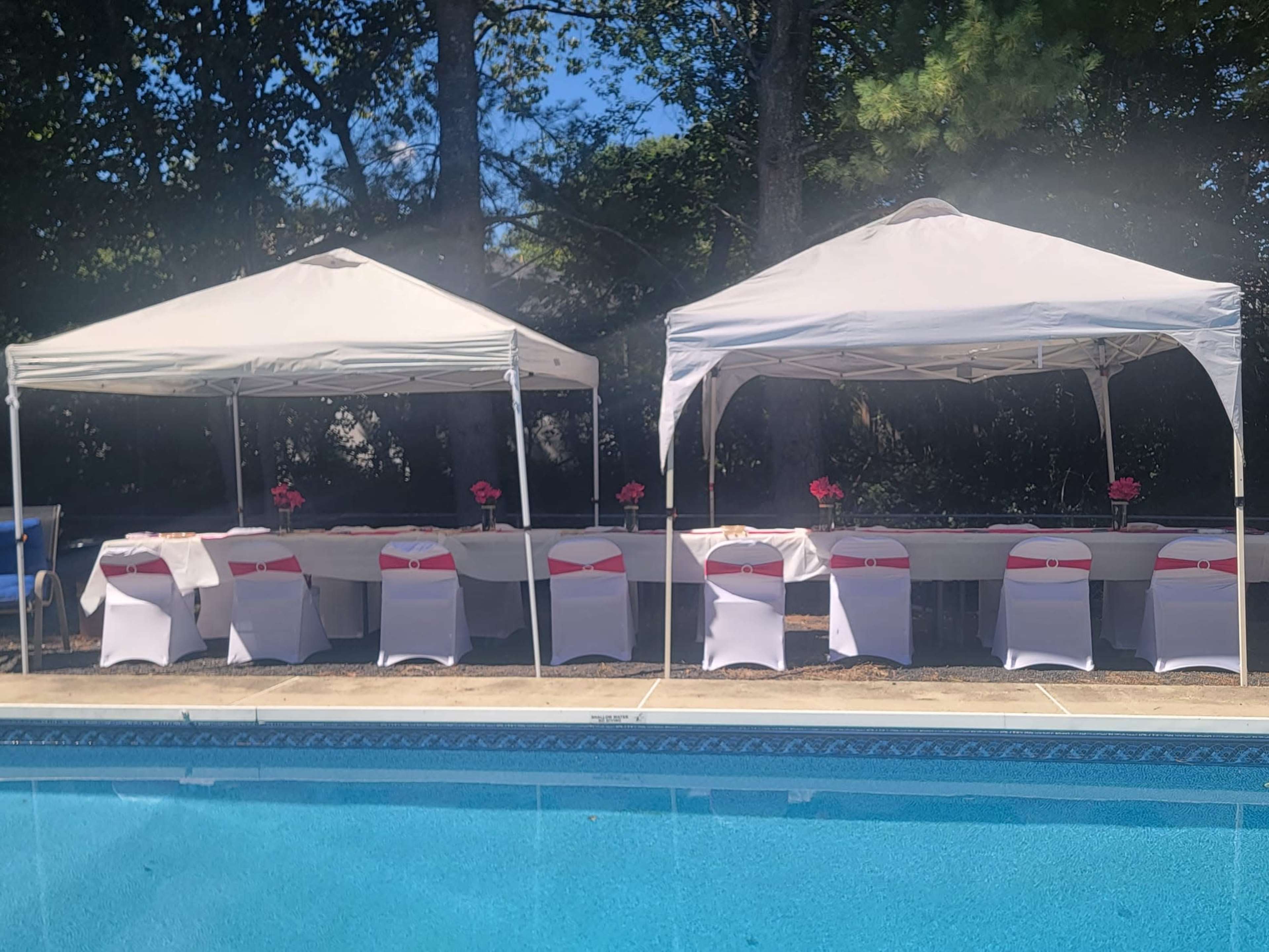 Two white canopies are set up beside a pool, with a long table decorated with pink flowers and chairs arranged in front of them.