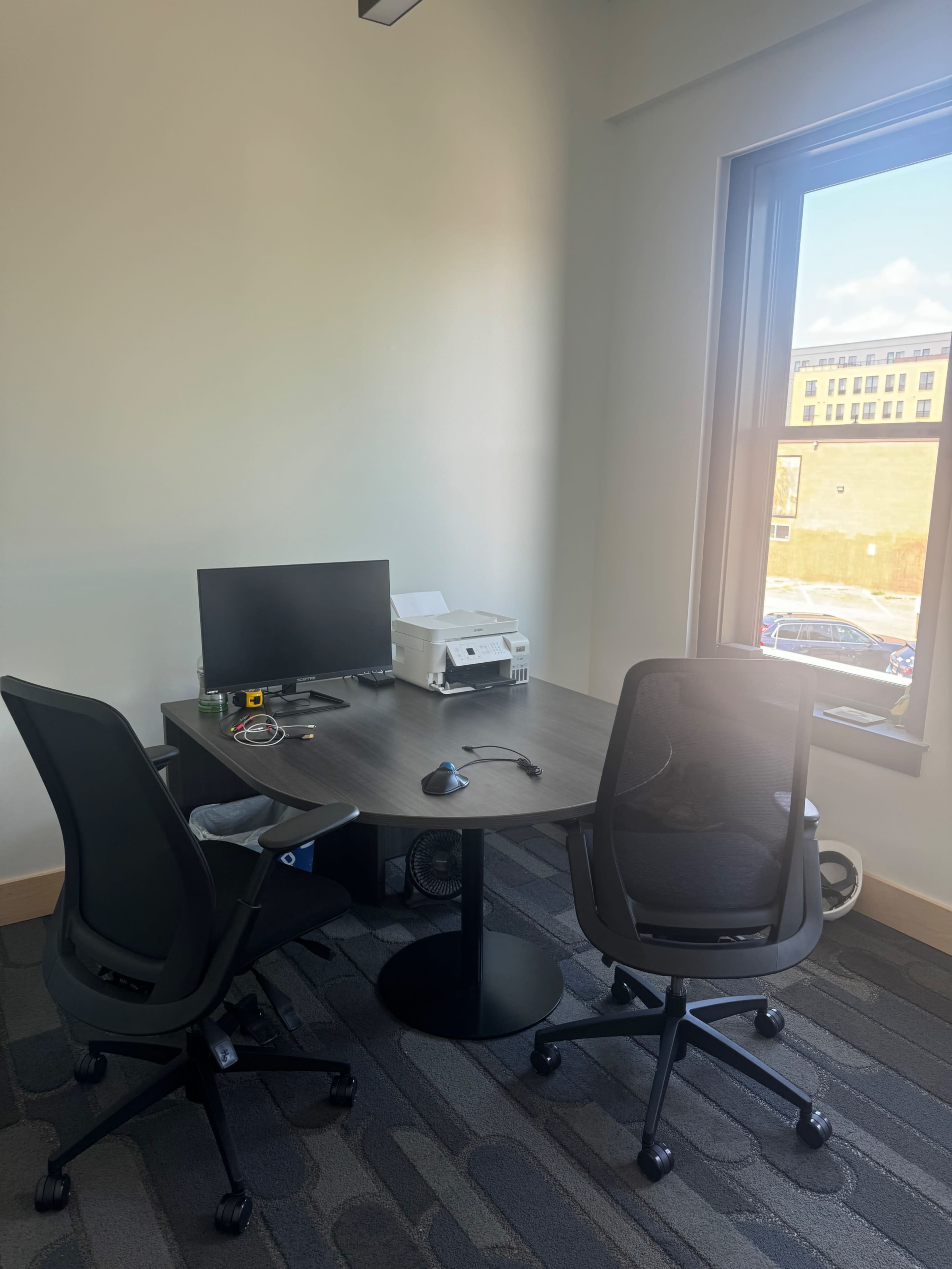 The image shows a small office space with a round table, two black chairs, a computer monitor, and a printer near a window.