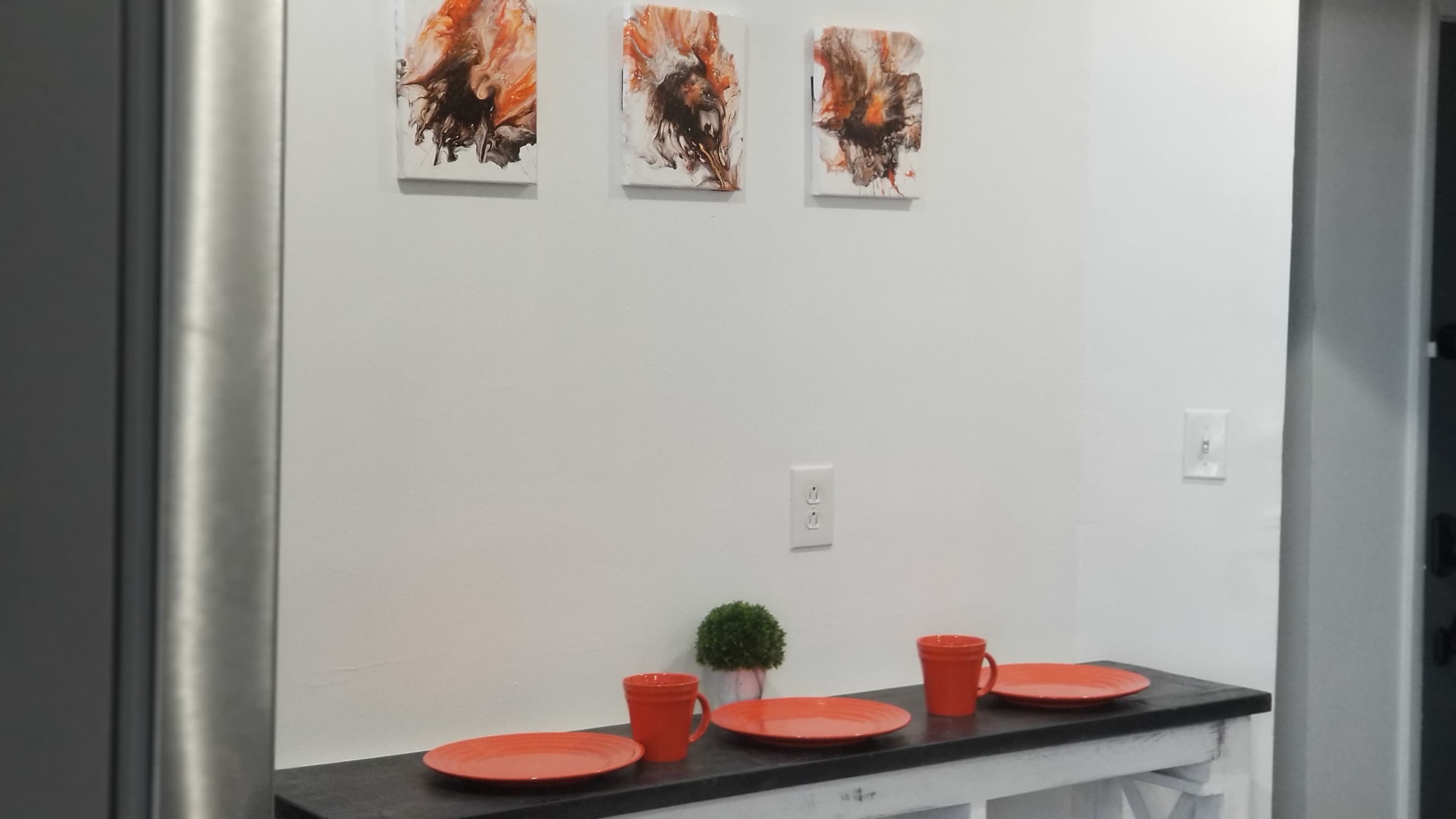 A white wall with three abstract art pieces in orange and black above a black countertop featuring four orange plates and two small orange pots with plants.