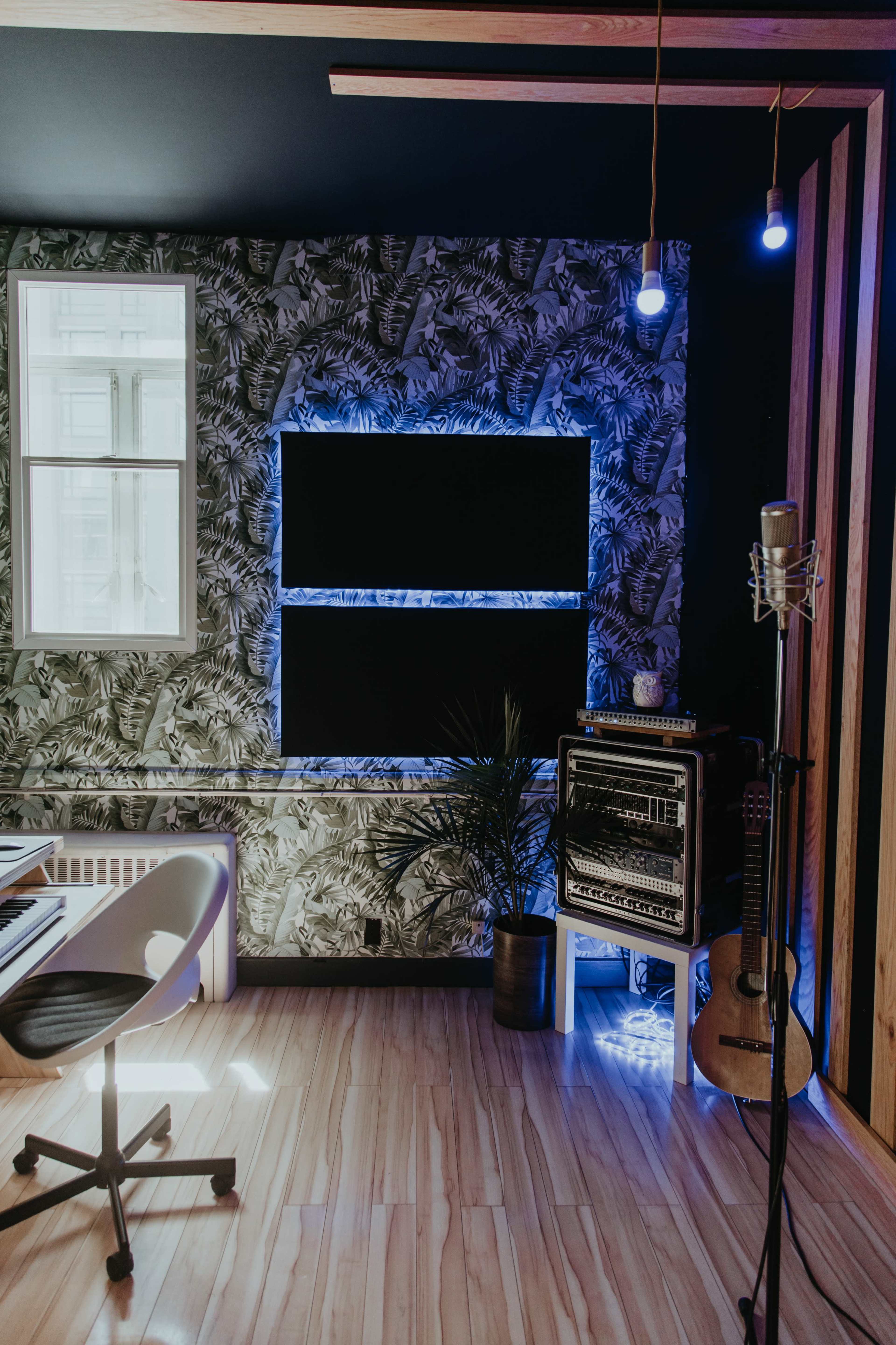 A modern recording studio with a plant, a guitar, and two screens illuminated by blue light against a patterned wall.