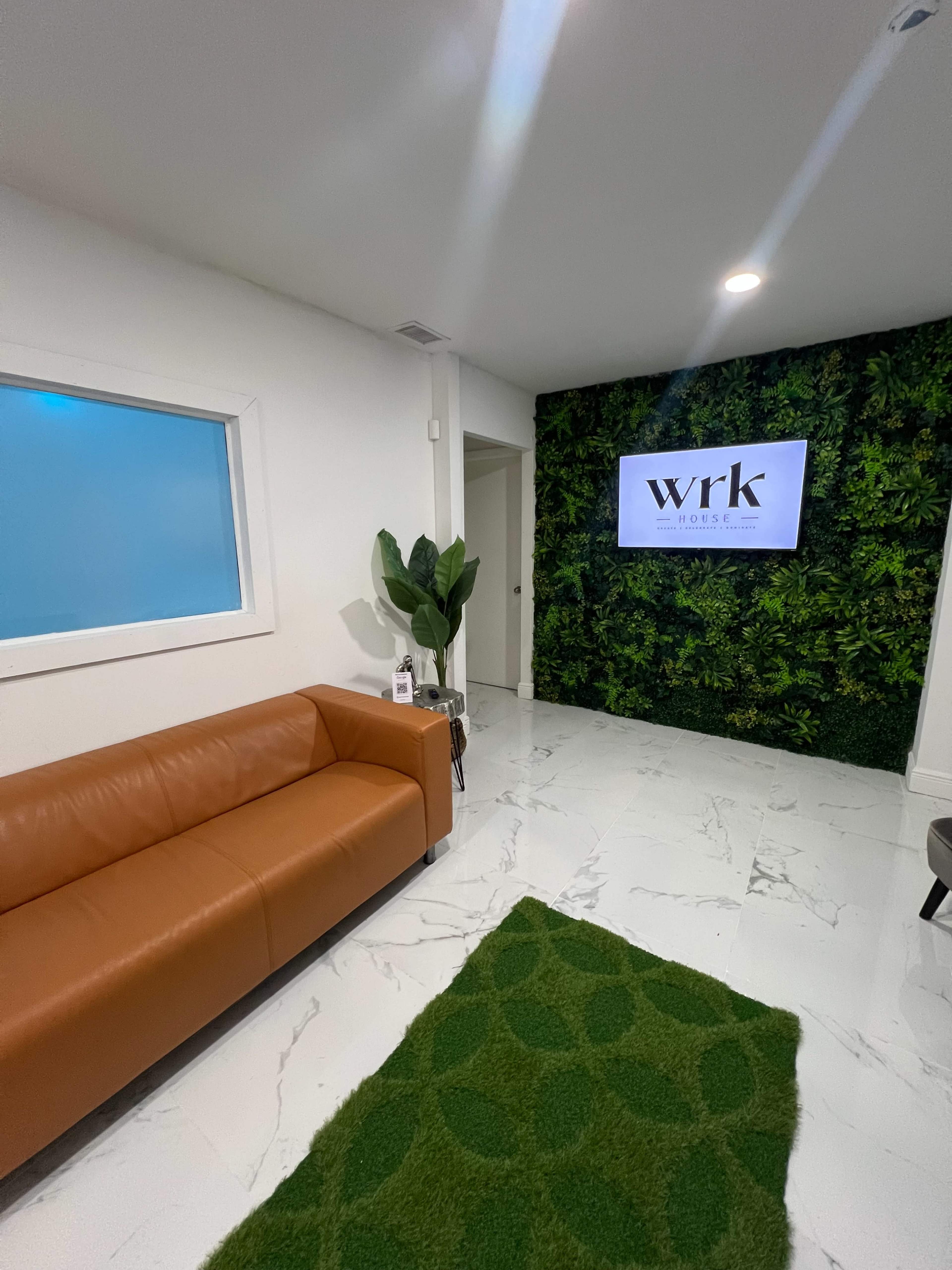 The image shows a modern waiting area with a brown leather couch, a green rug, a plant, and a wall adorned with greenery featuring a television screen displaying the letters "wrk."