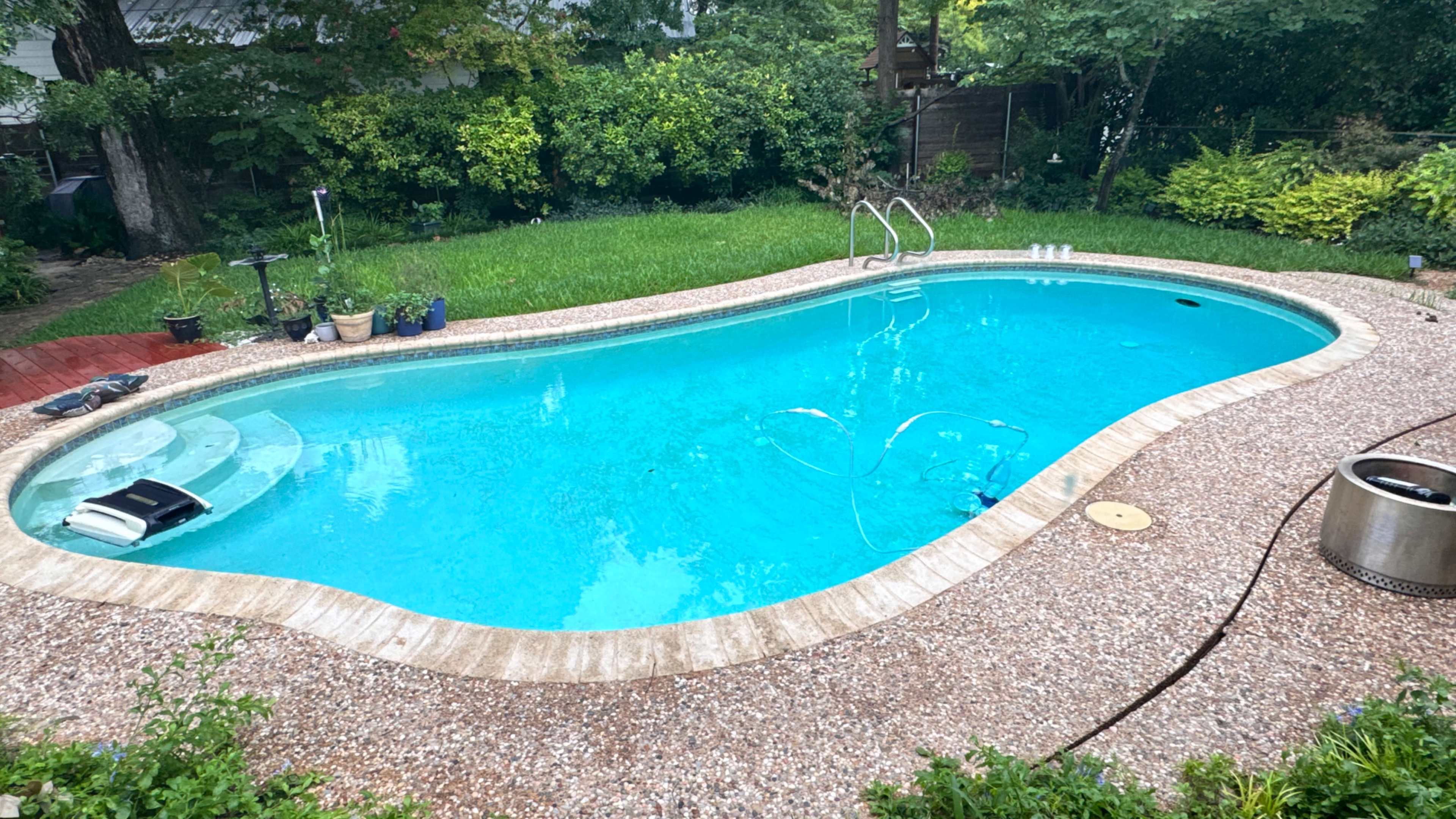 The image shows an oval swimming pool surrounded by a pebbled deck and greenery in the background.