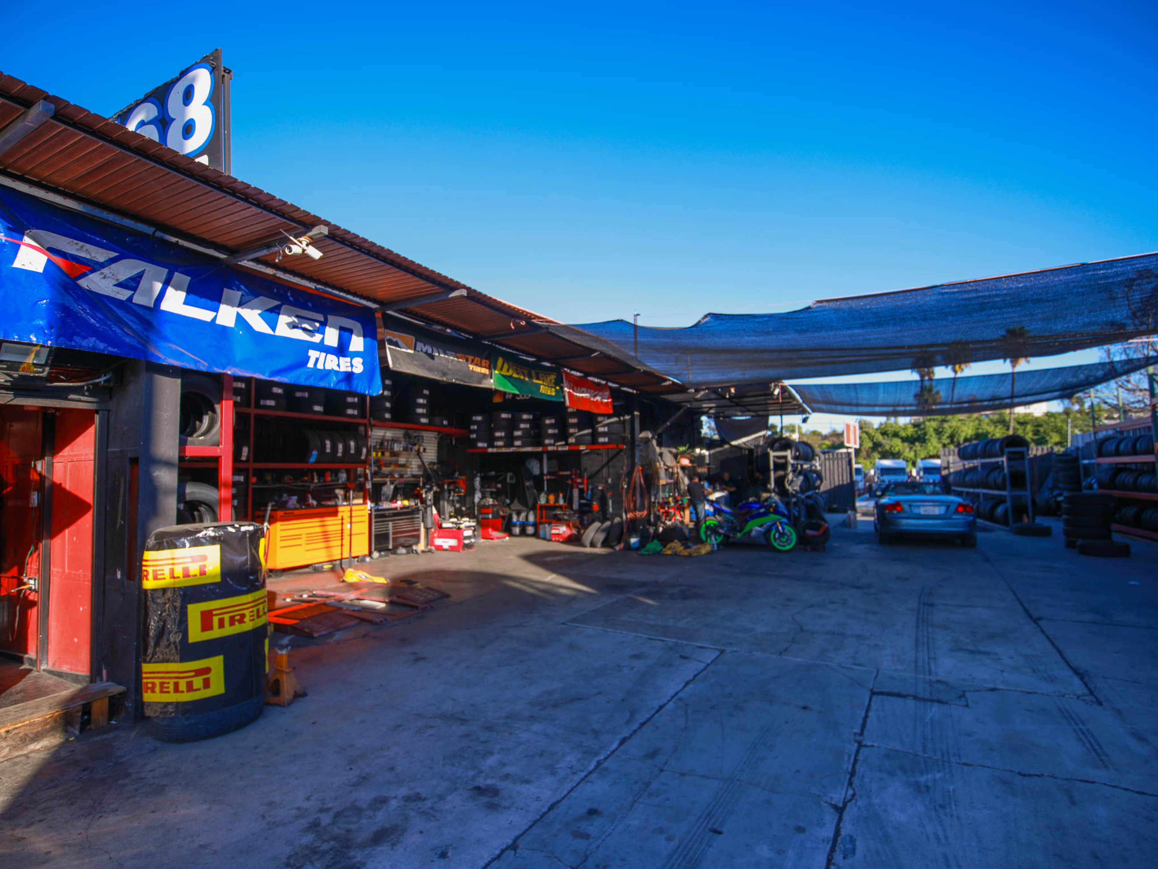The image shows a tire shop with various tire brands displayed, including Falken and Pirelli, alongside tools and equipment in a garage area under clear blue skies.