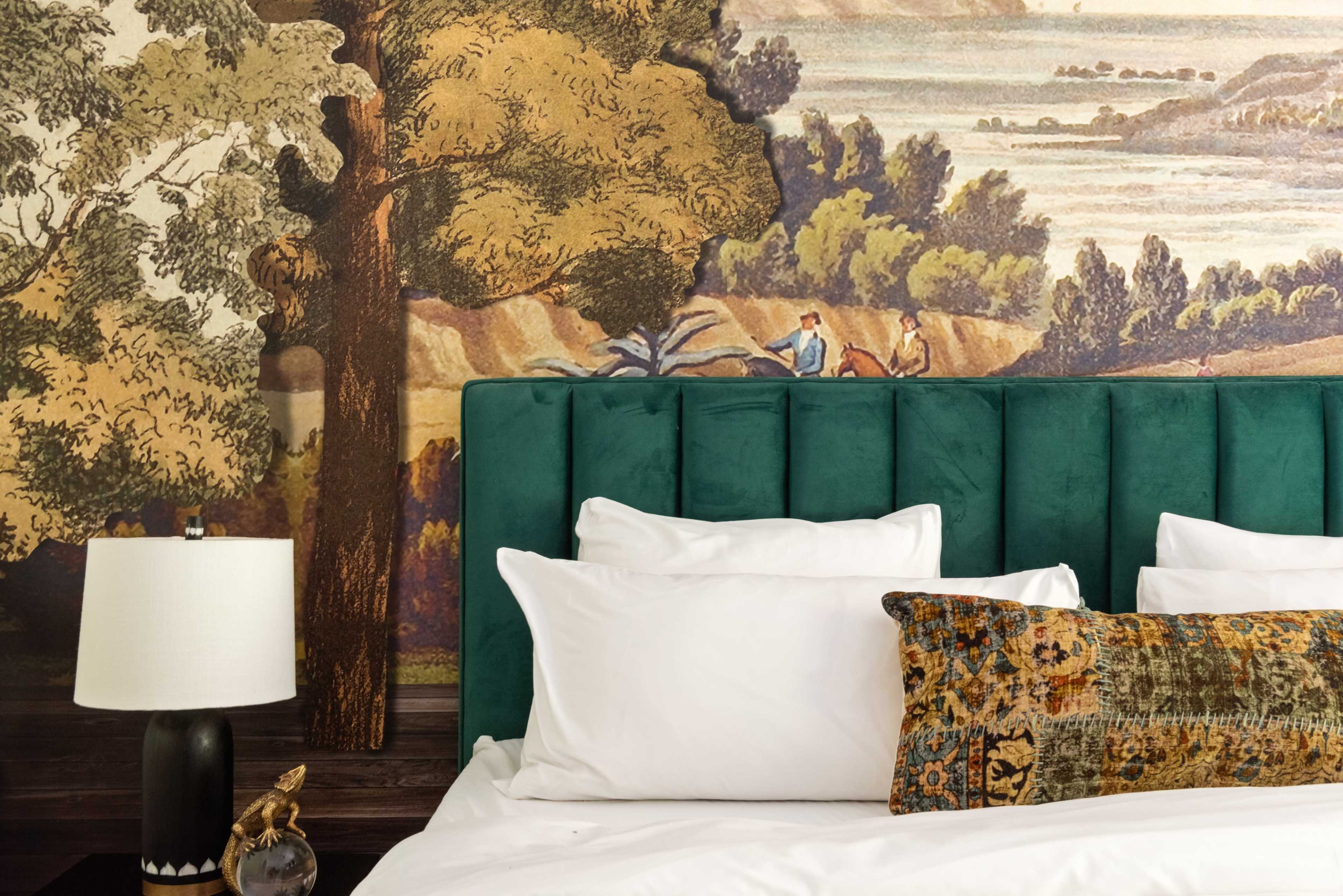 The image features a stylish bedroom with a deep green upholstered headboard, white bedding, and a decorative throw pillow, set against a large mural depicting a landscape scene.
