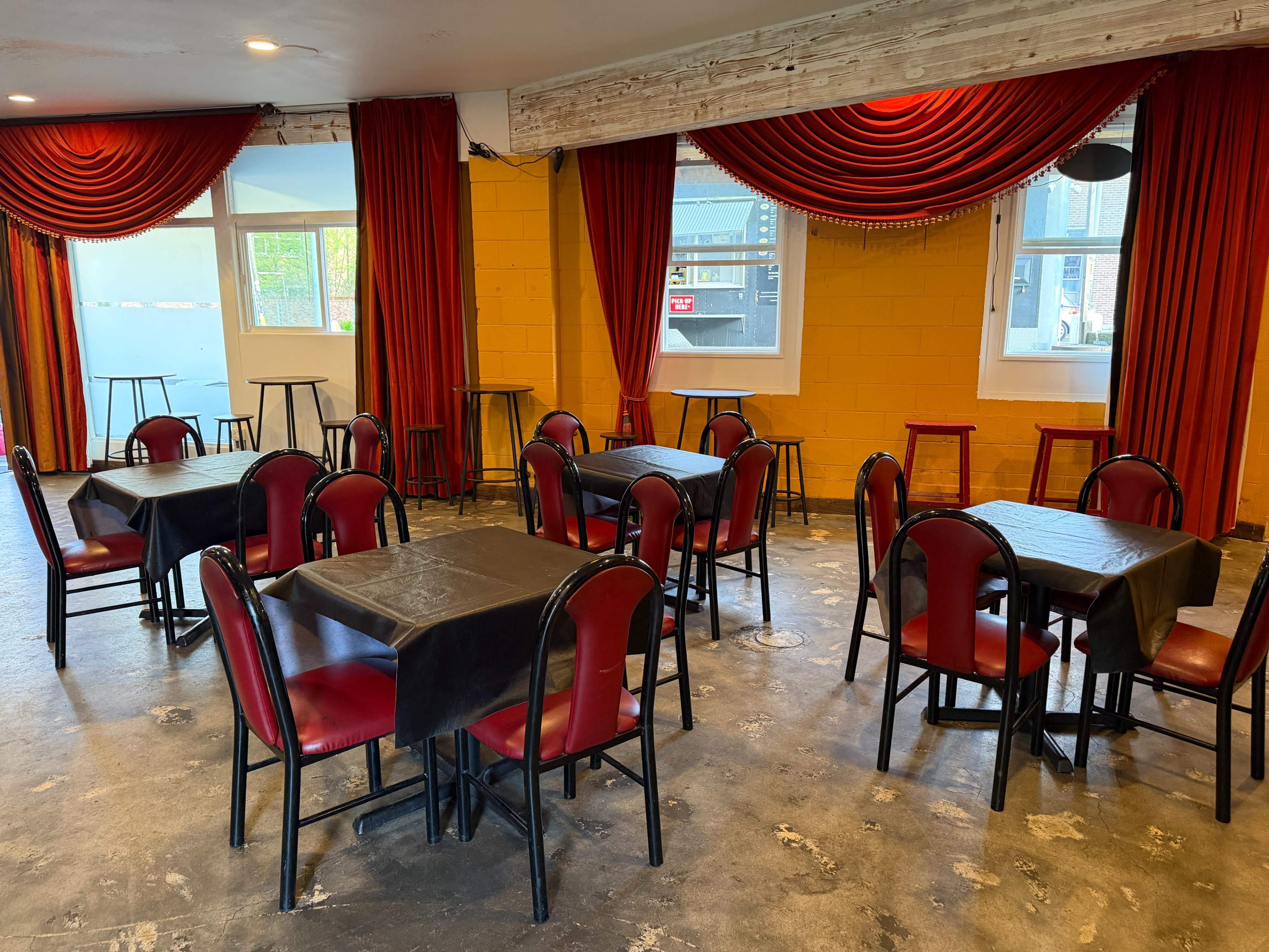 The image shows a restaurant interior with several tables and chairs arranged under red curtains and large windows.