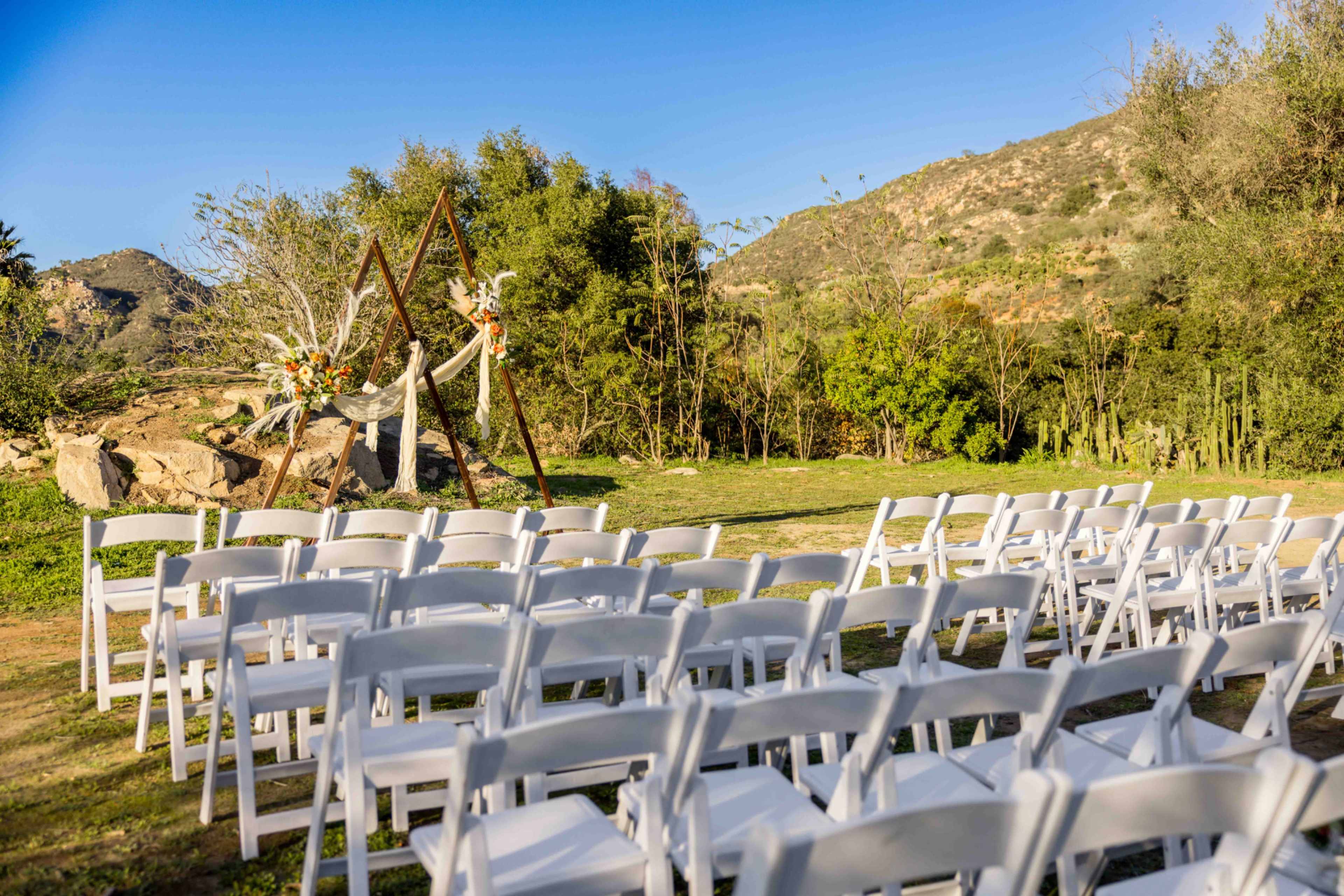 The image shows an outdoor wedding setting with rows of white chairs facing a floral-decorated arch set against a mountainous backdrop.