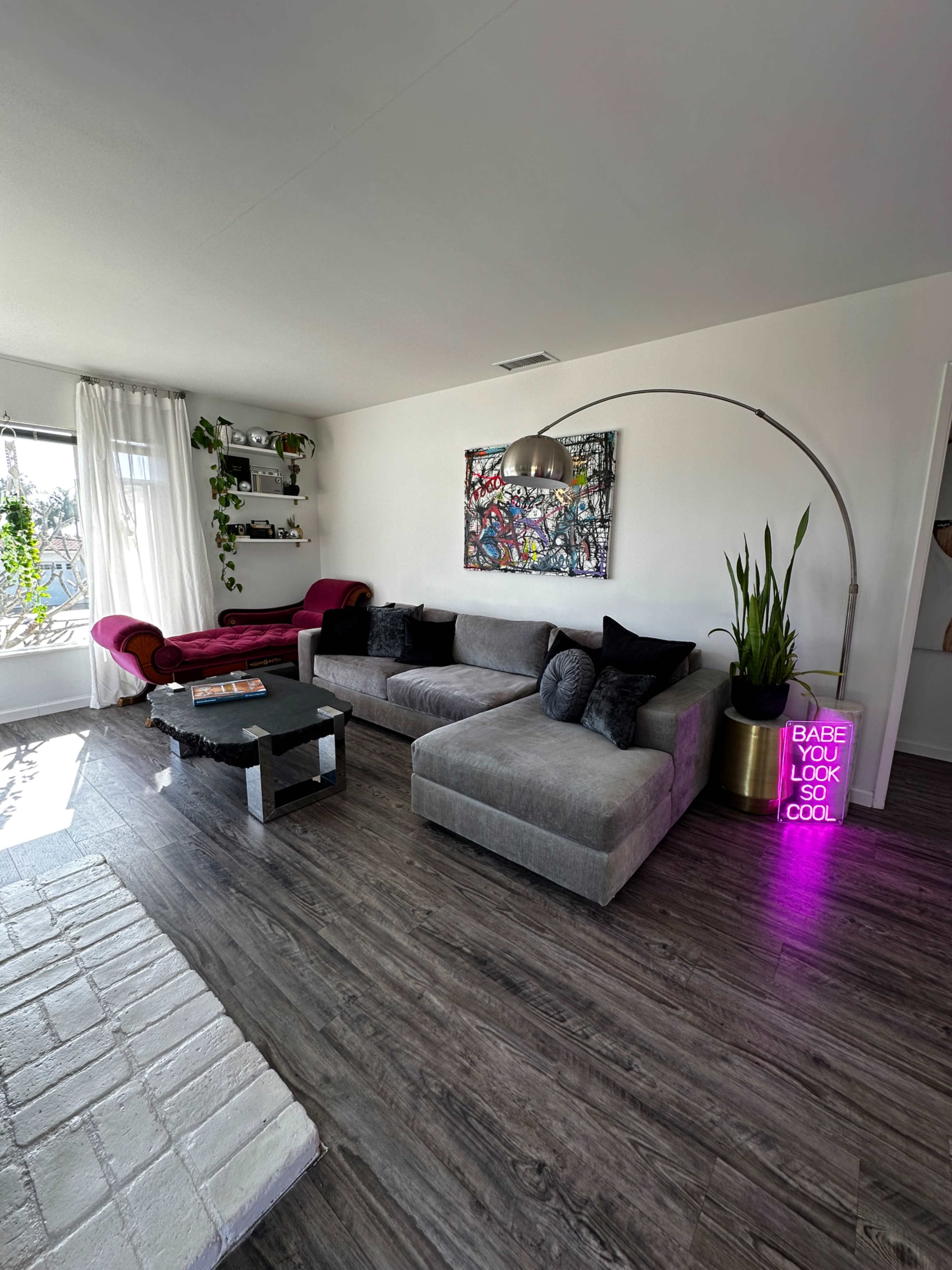 The living room features a large gray sectional sofa, a black coffee table, and a colorful wall art piece, with a neon sign and potted plant in the corner.