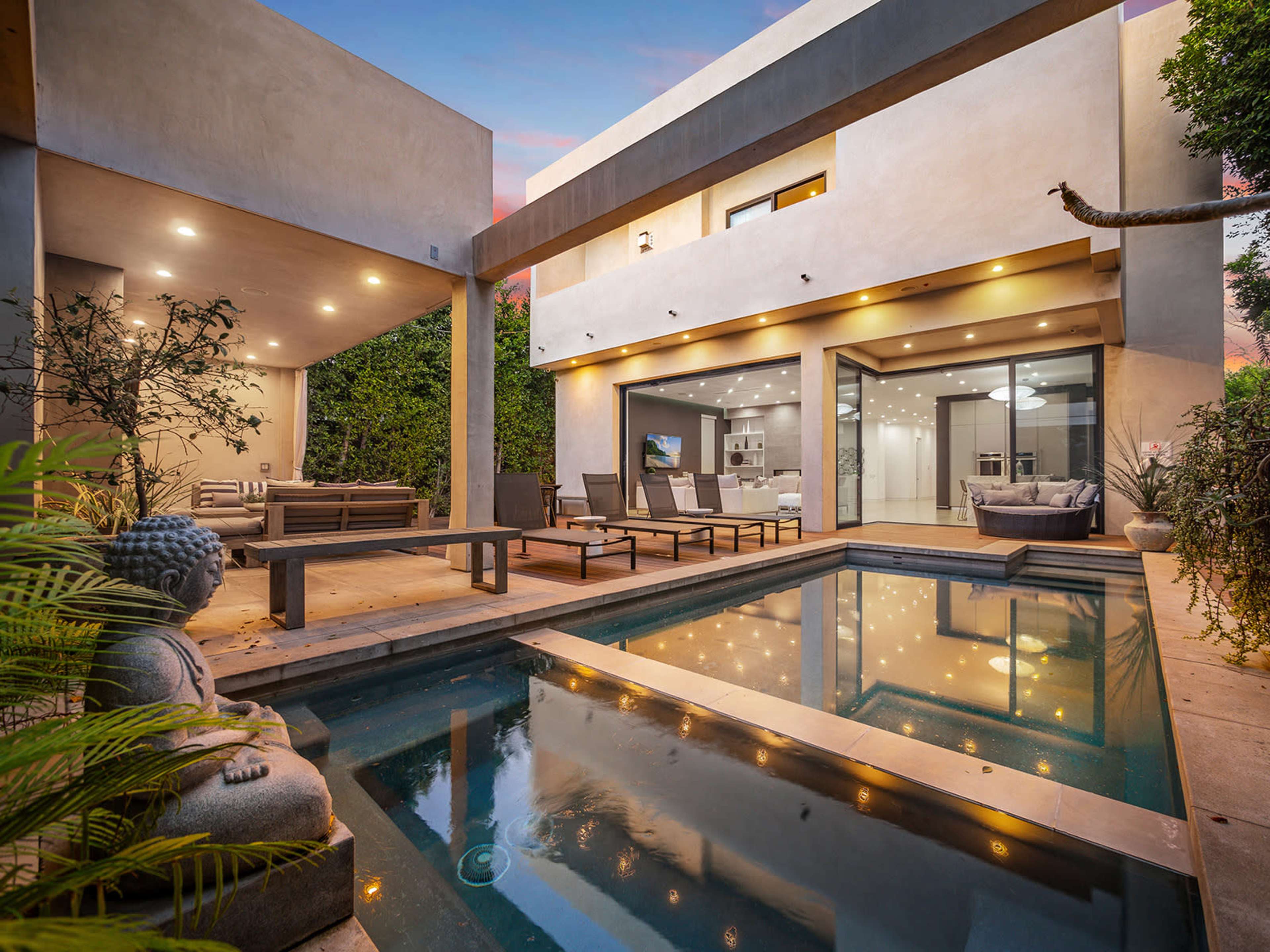 A modern outdoor space featuring a swimming pool, lounge chairs, and a statue, with an adjacent house illuminated by soft lighting.