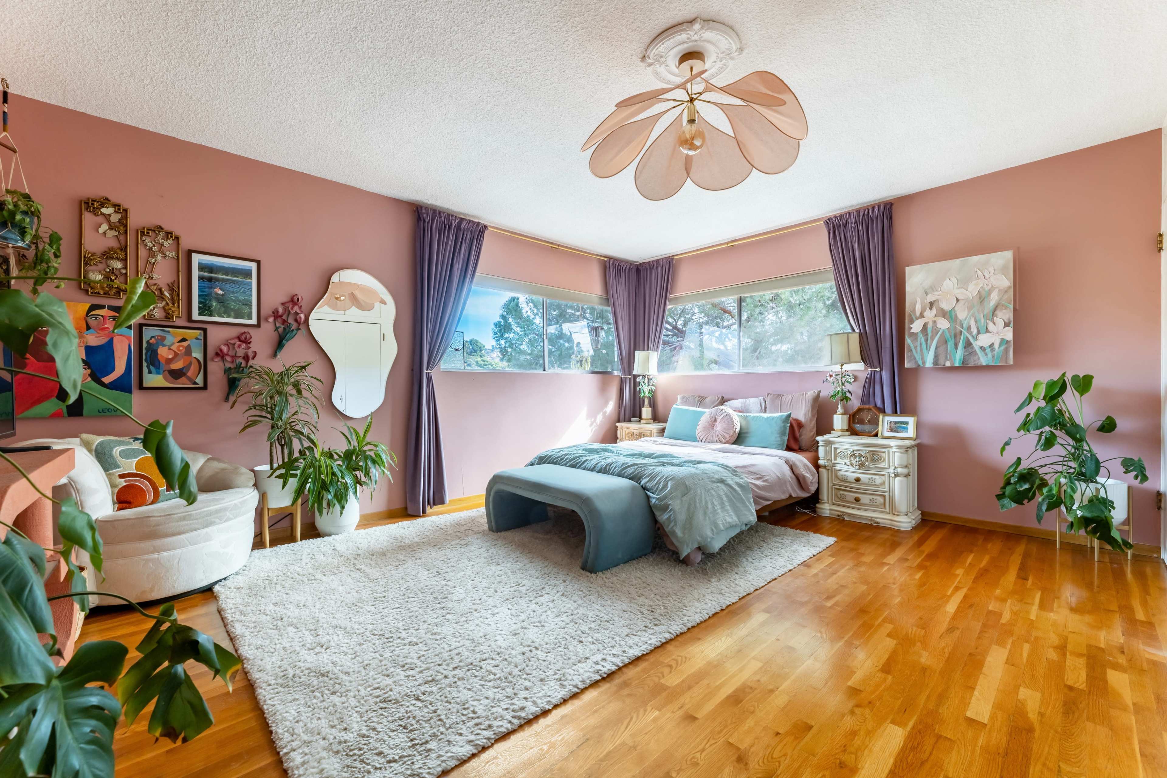 The image shows a cozy bedroom featuring a large bed, a plush rug, houseplants, and a mix of colorful decor on the walls.