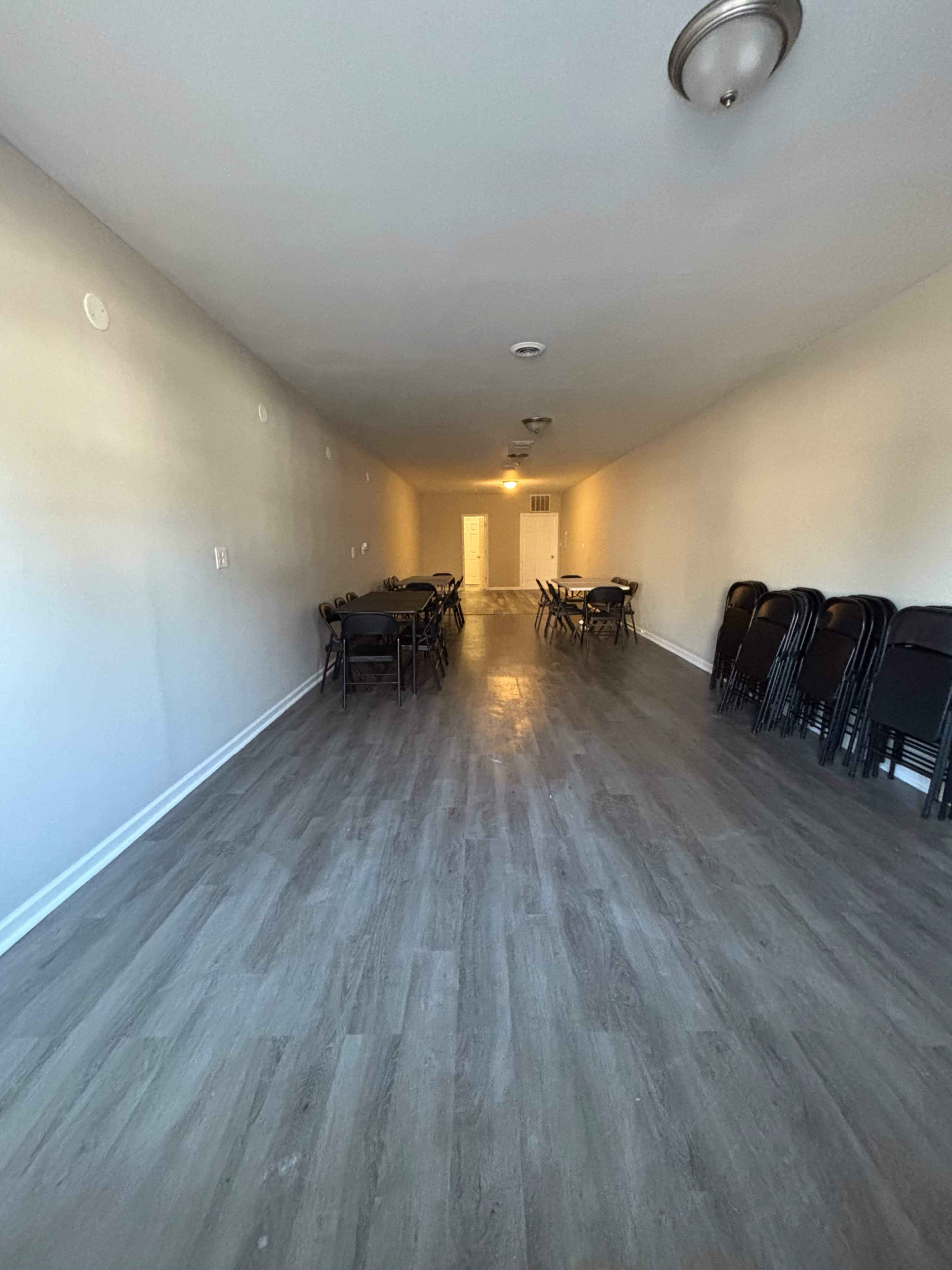 The image shows a long, narrow room with a smooth floor, featuring several folding tables and chairs arranged along the sides.