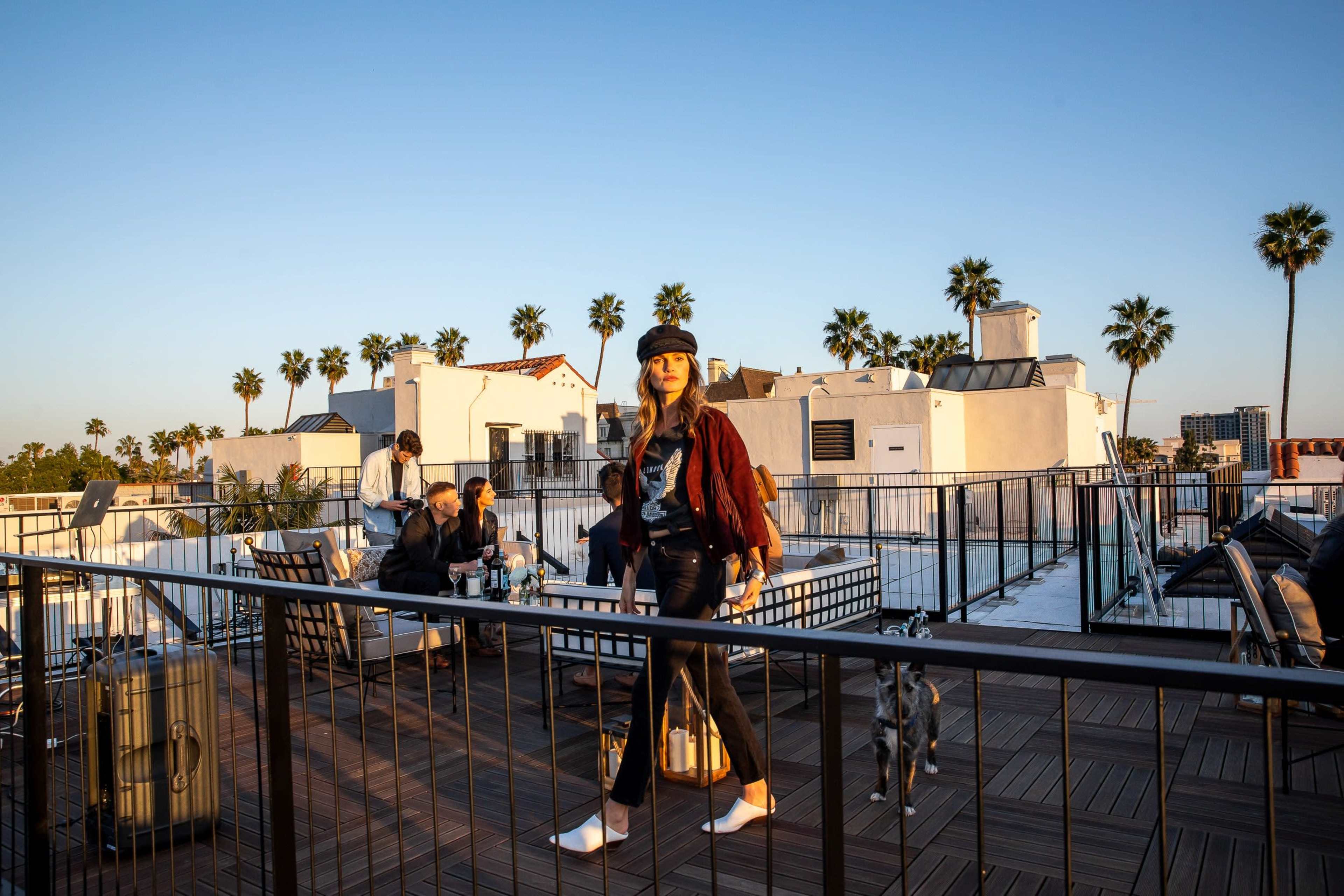 A woman walks on a rooftop terrace with palm trees and people seated in the background.