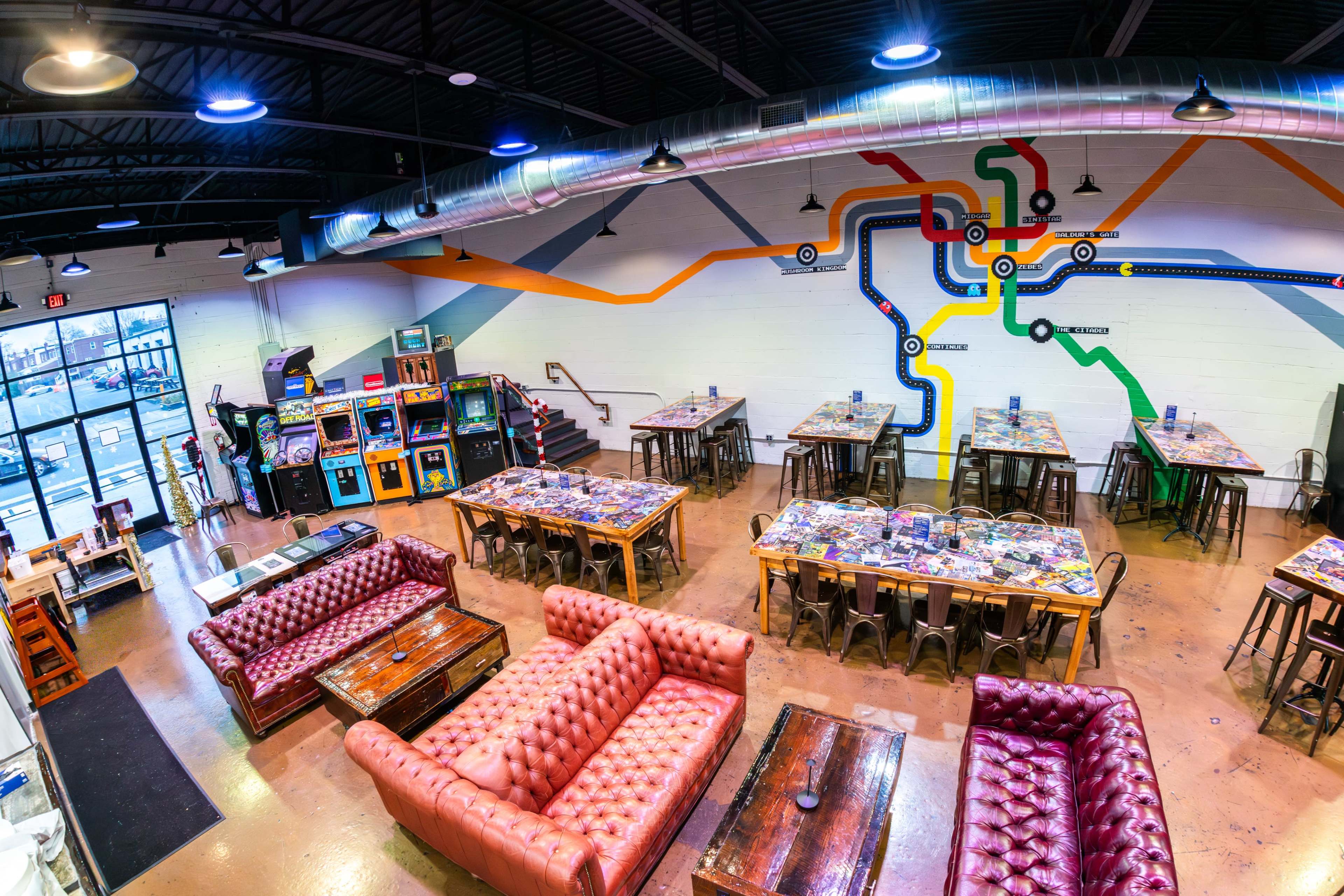 The image shows a vibrant, open dining area with red tufted couches, wooden tables, and an arcade game corner, featuring a colorful mural on the wall.