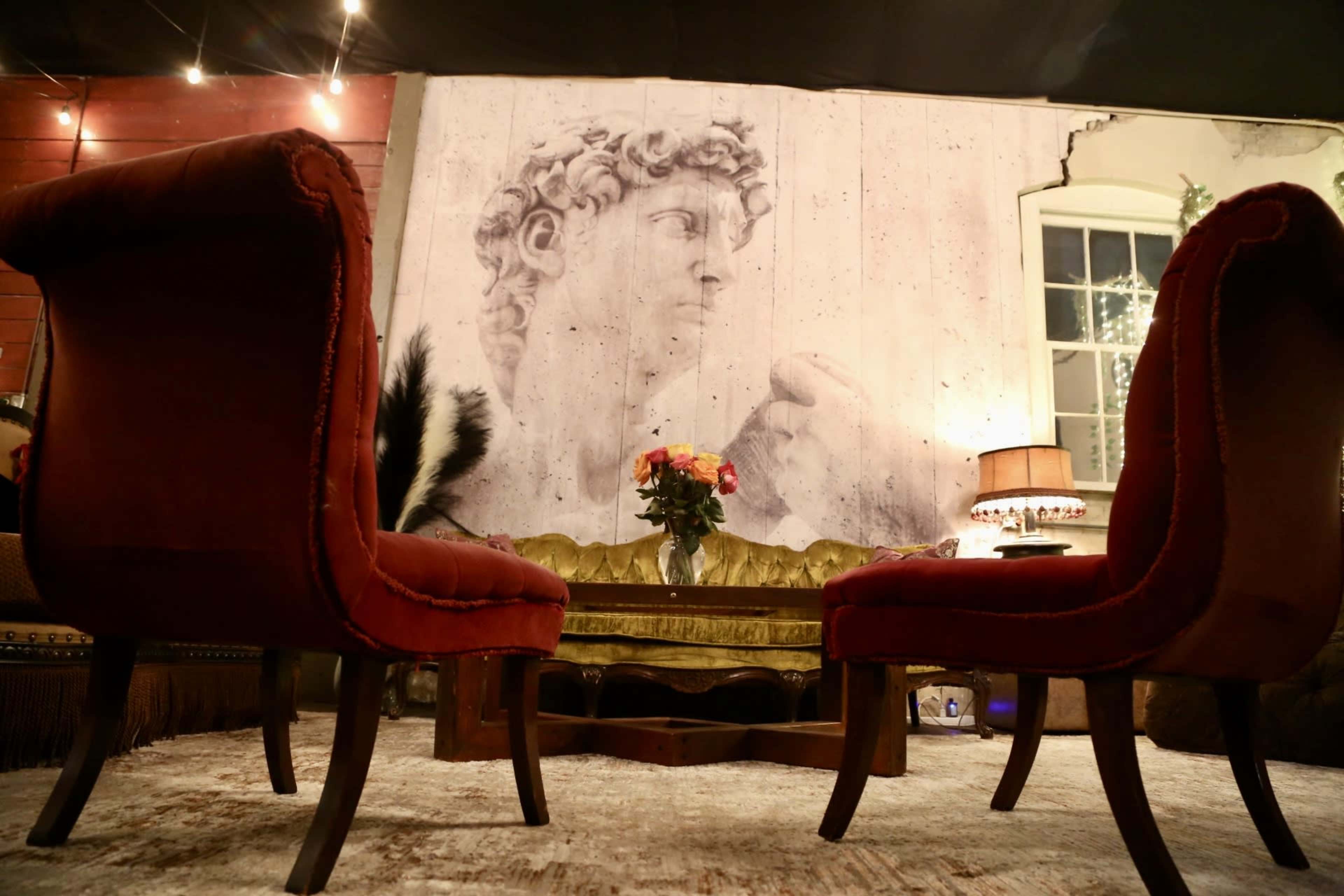 The image shows two red armchairs facing a wooden coffee table, with a large mural of a classical statue on the wall behind them.