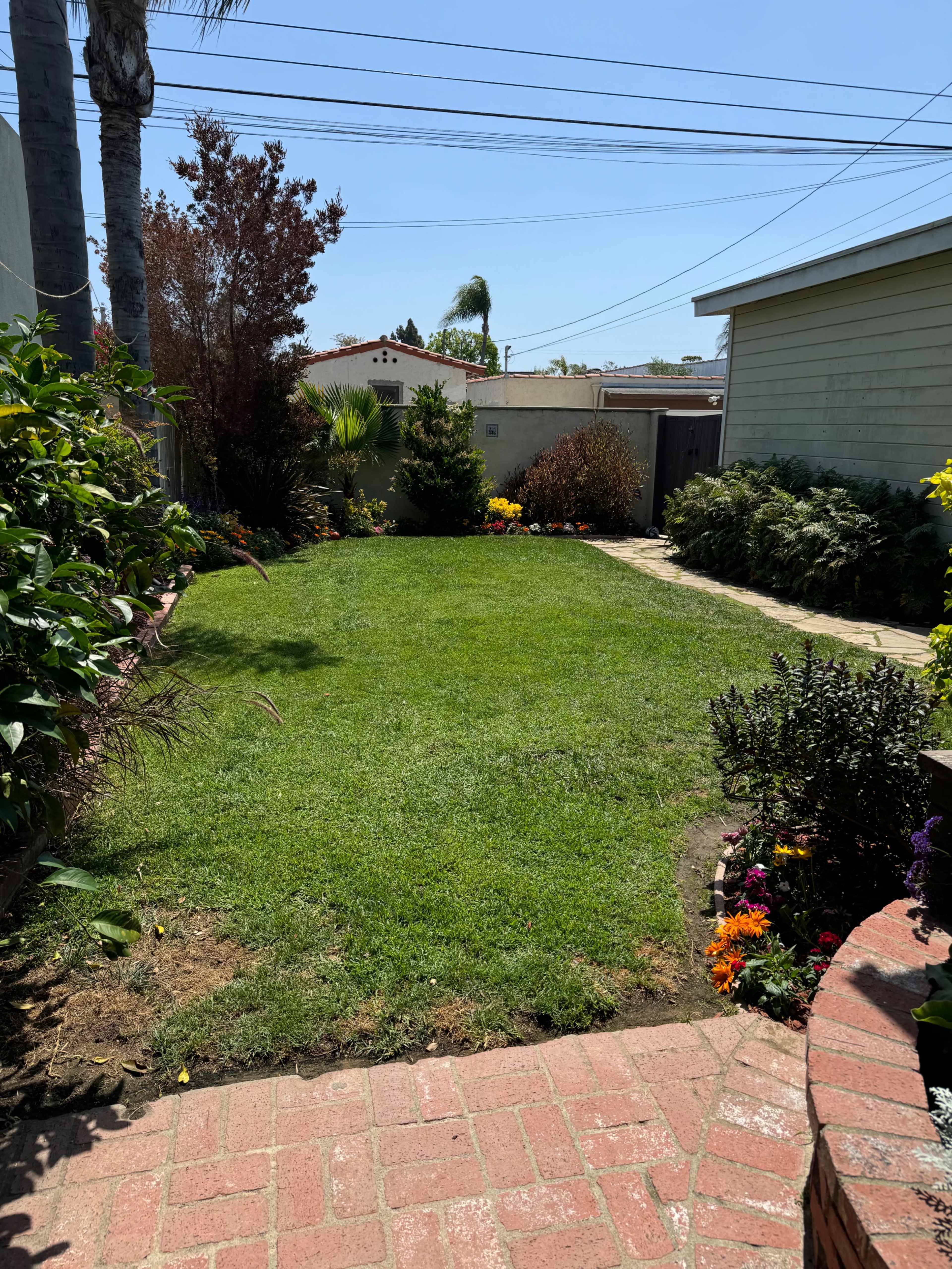 A well-maintained back lawn is bordered by flower beds and leads to a pathway beside a house.