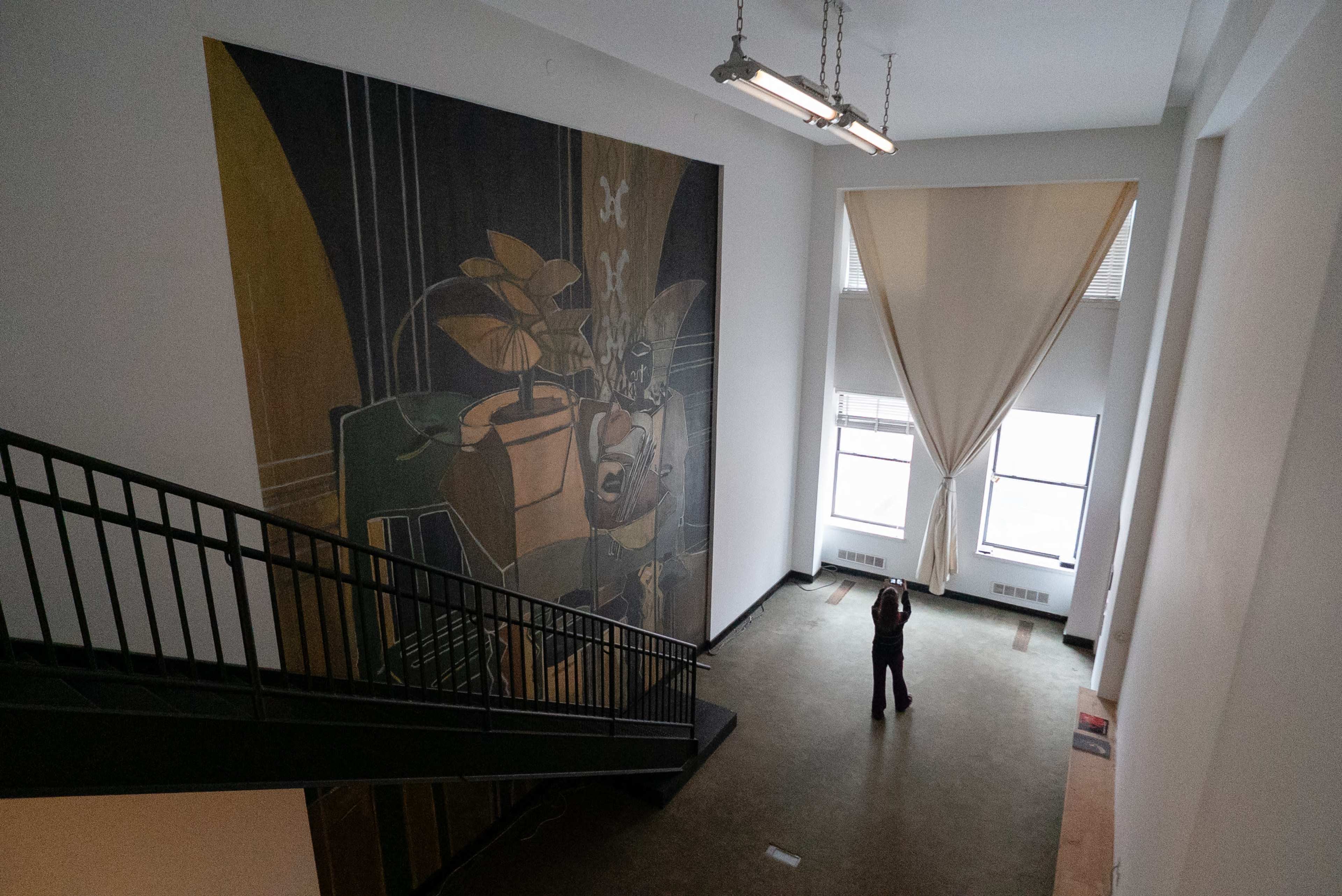 A person stands on a lower floor, looking up at a large mural on the wall, with a staircase in the foreground and tall windows nearby.