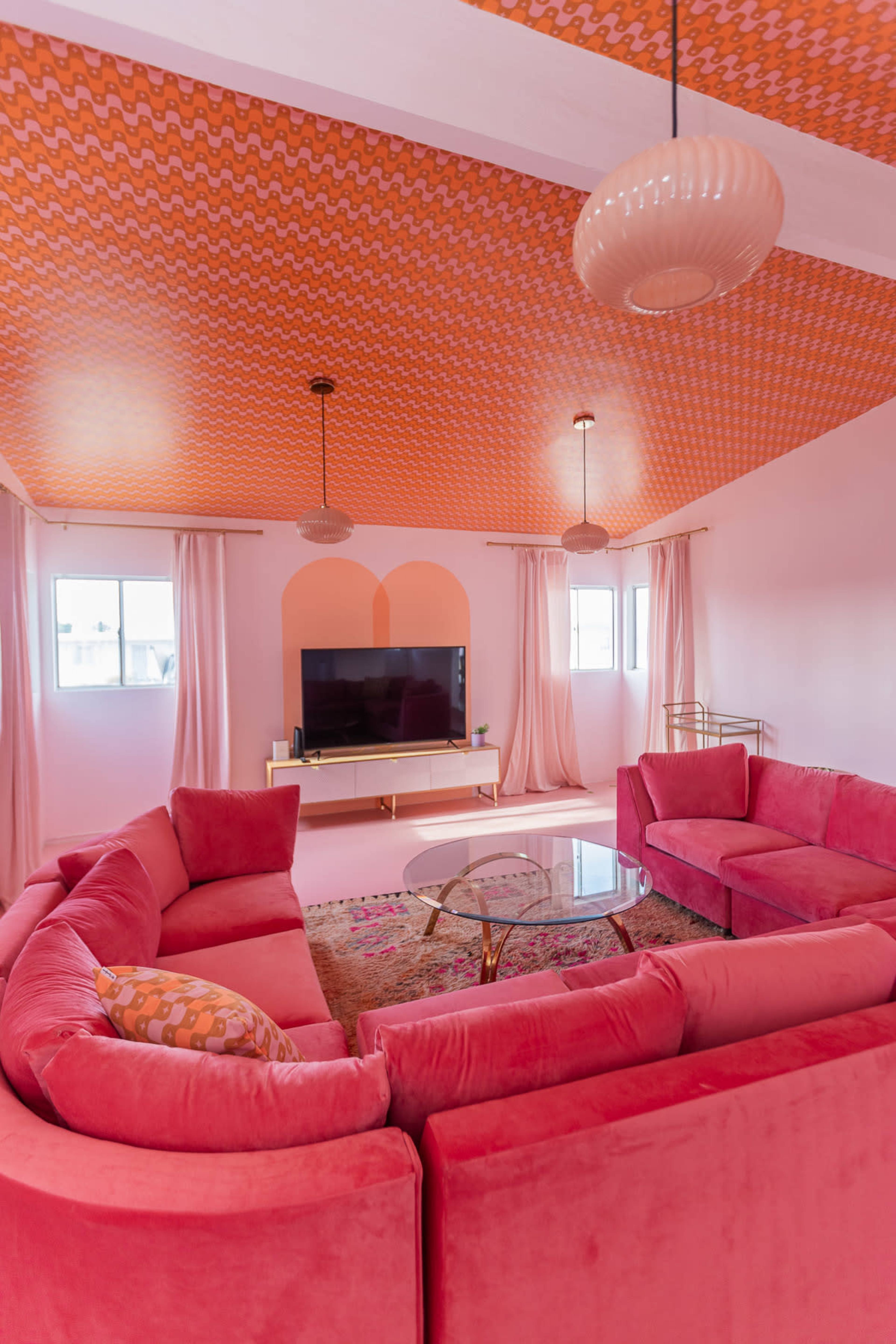 The room features a pink sectional sofa arranged around a glass coffee table, with a colorful ceiling and a television set on a gold console against the wall.