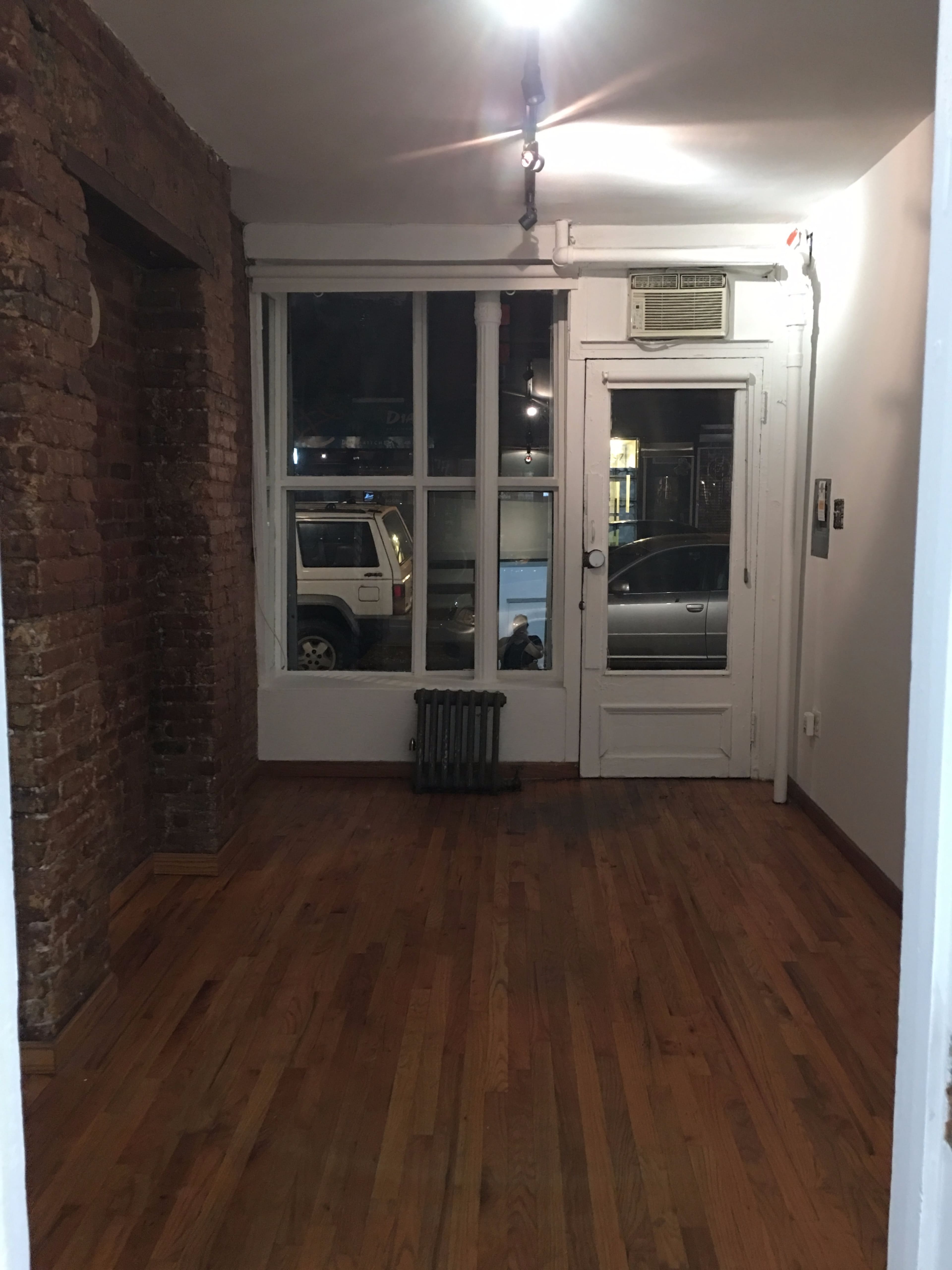 The image shows a sparsely furnished room with exposed brick walls, a radiator, a window revealing a street view, and a door leading outside.