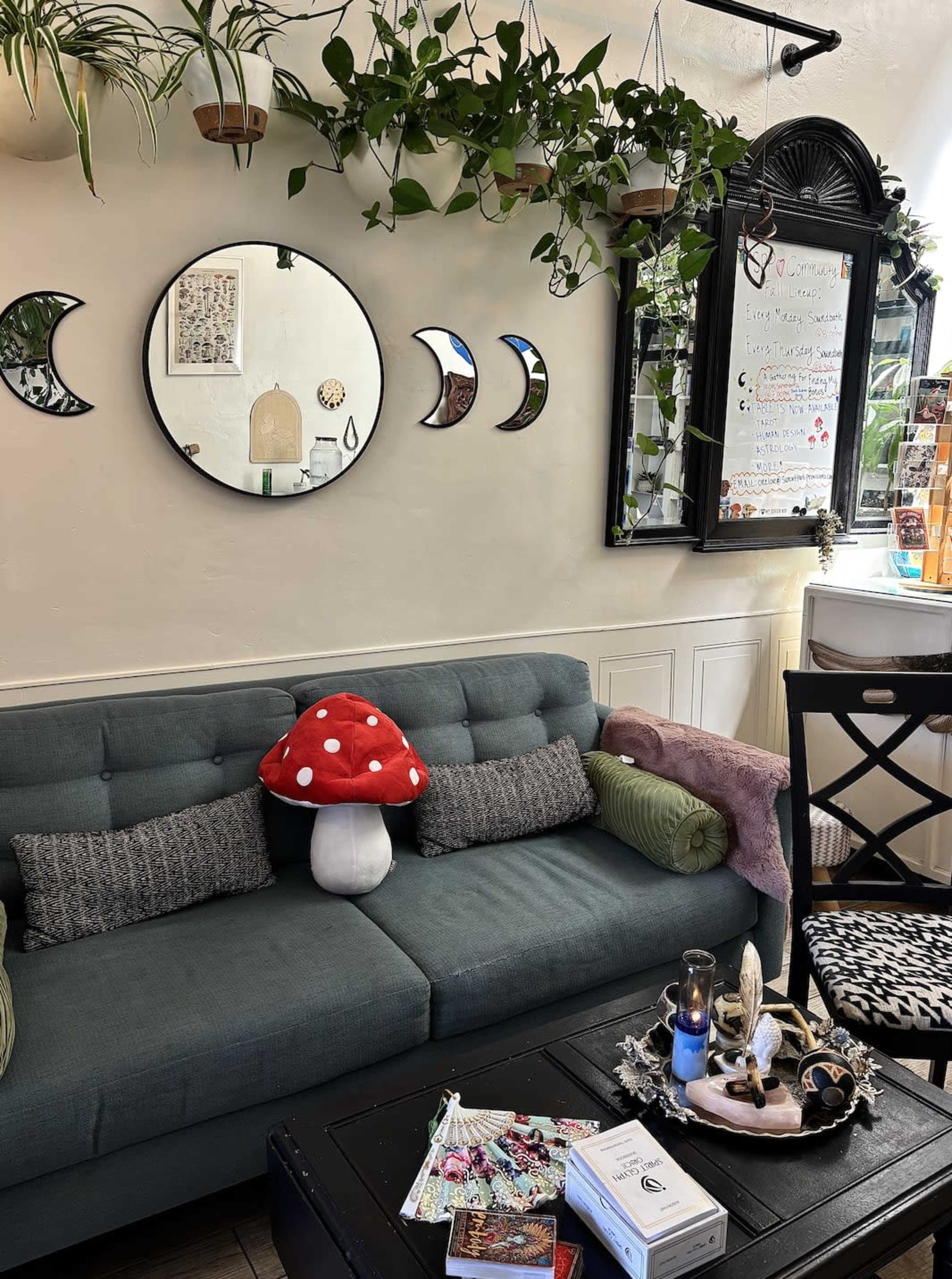 The image shows a cozy indoor sitting area featuring a dark green couch with decorative pillows, a large red-and-white mushroom plush, a round mirror on the wall, and various potted plants hanging above.