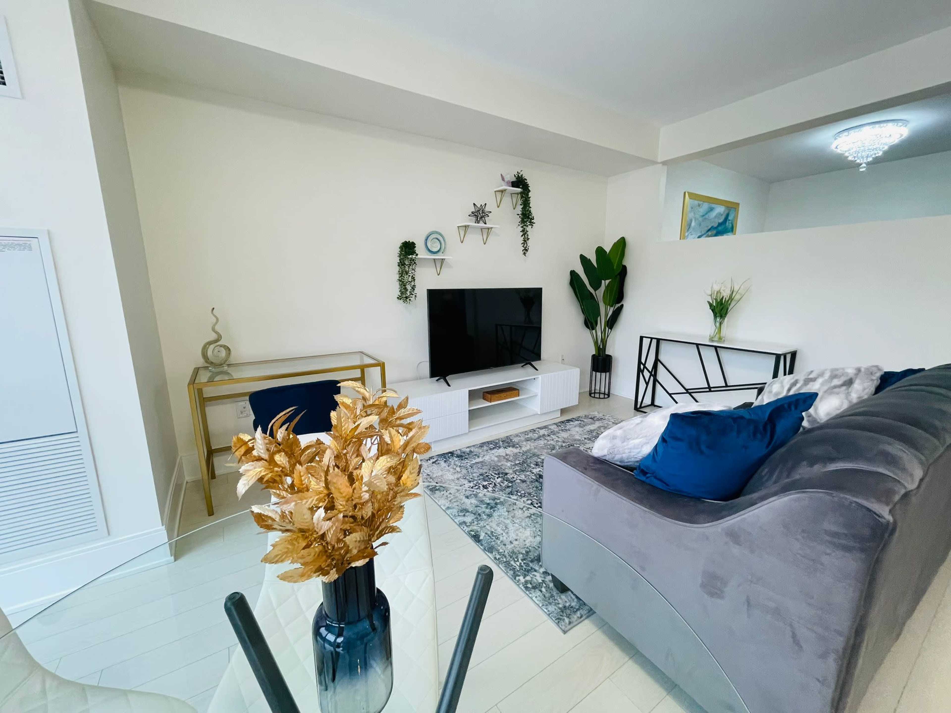A modern living room with a television mounted on the wall, a light-colored sofa, a glass coffee table, and decorative plants.